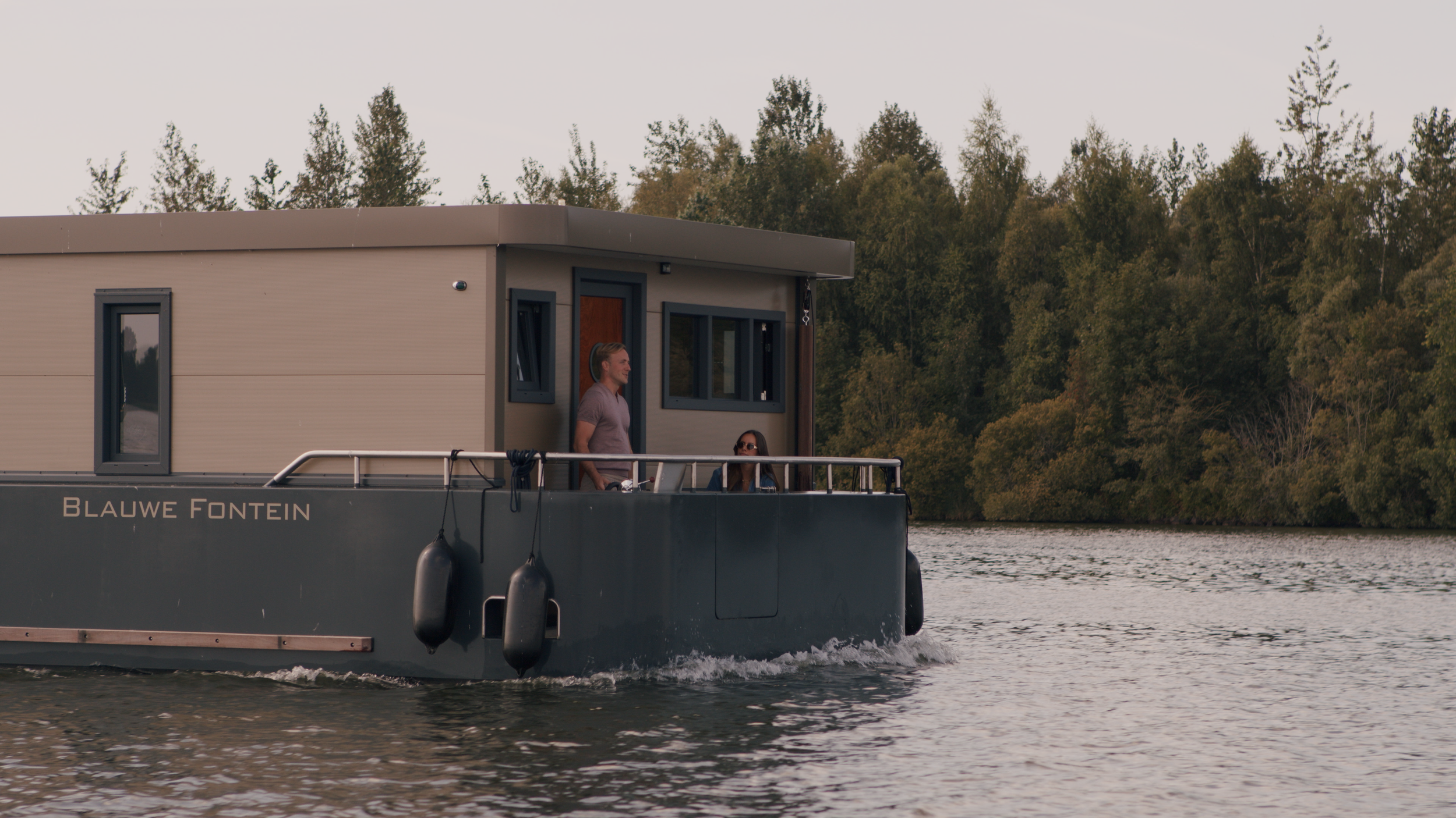 Houseboat named 'Blauwe Fontein' on a river with two people on the deck, surrounded by trees.