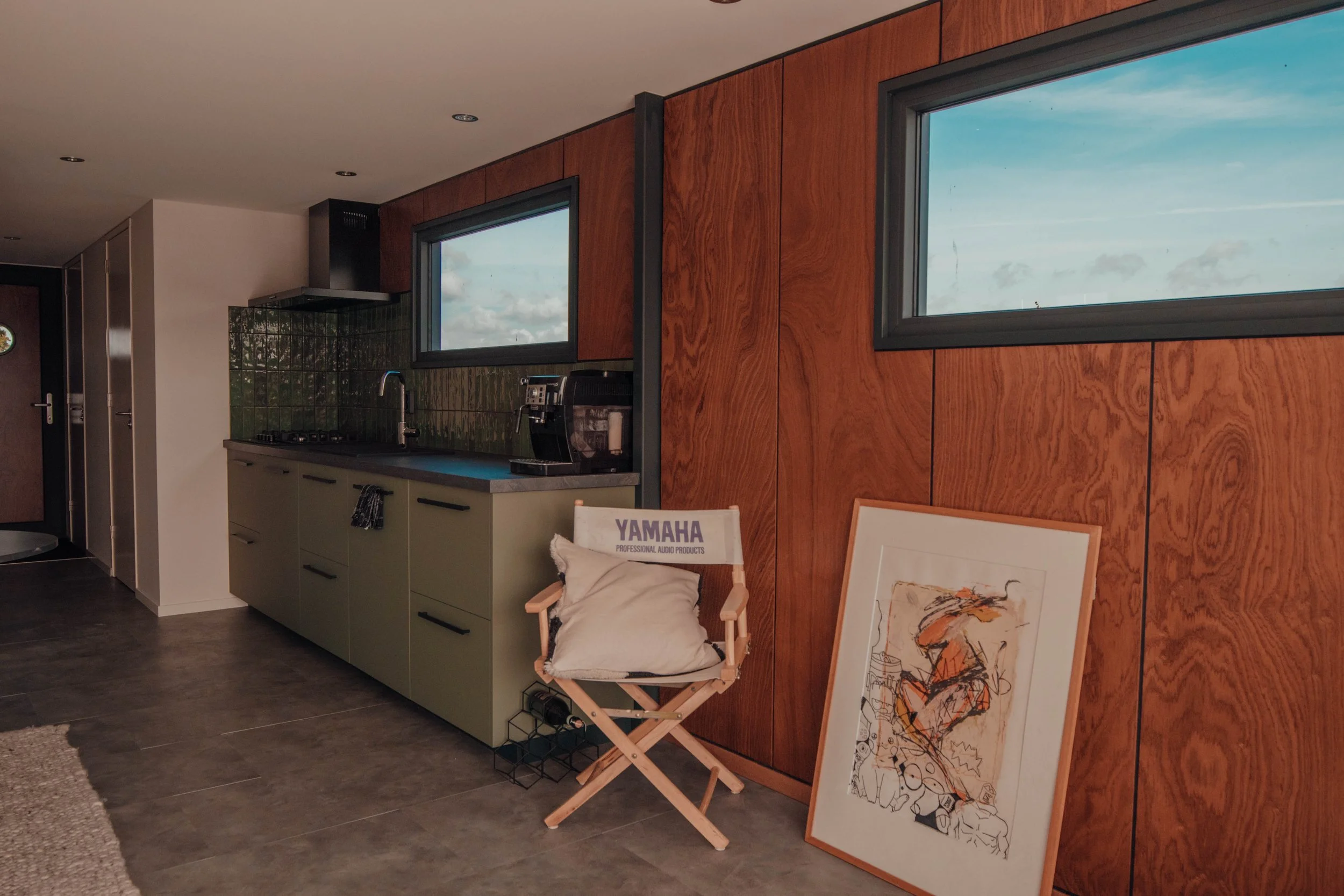 Modern kitchen interior with green cabinets, tiled backsplash, and wall art. A chair with a cushion is placed beside the counter. Two rectangular windows provide natural light.