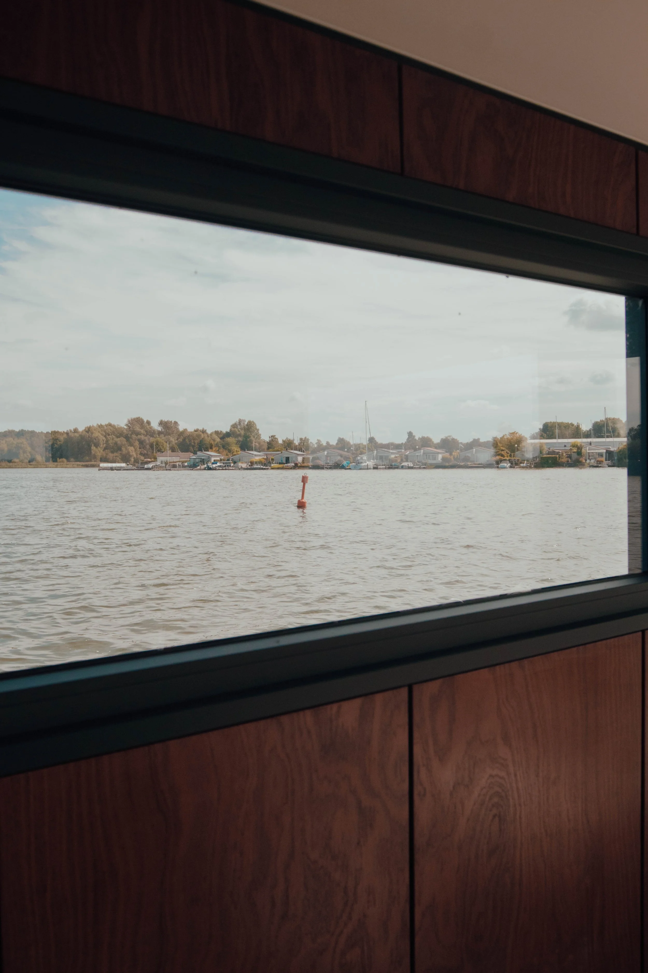 View through a boat window showing a river with a buoy, distant shoreline, and trees, framed by wood paneling.