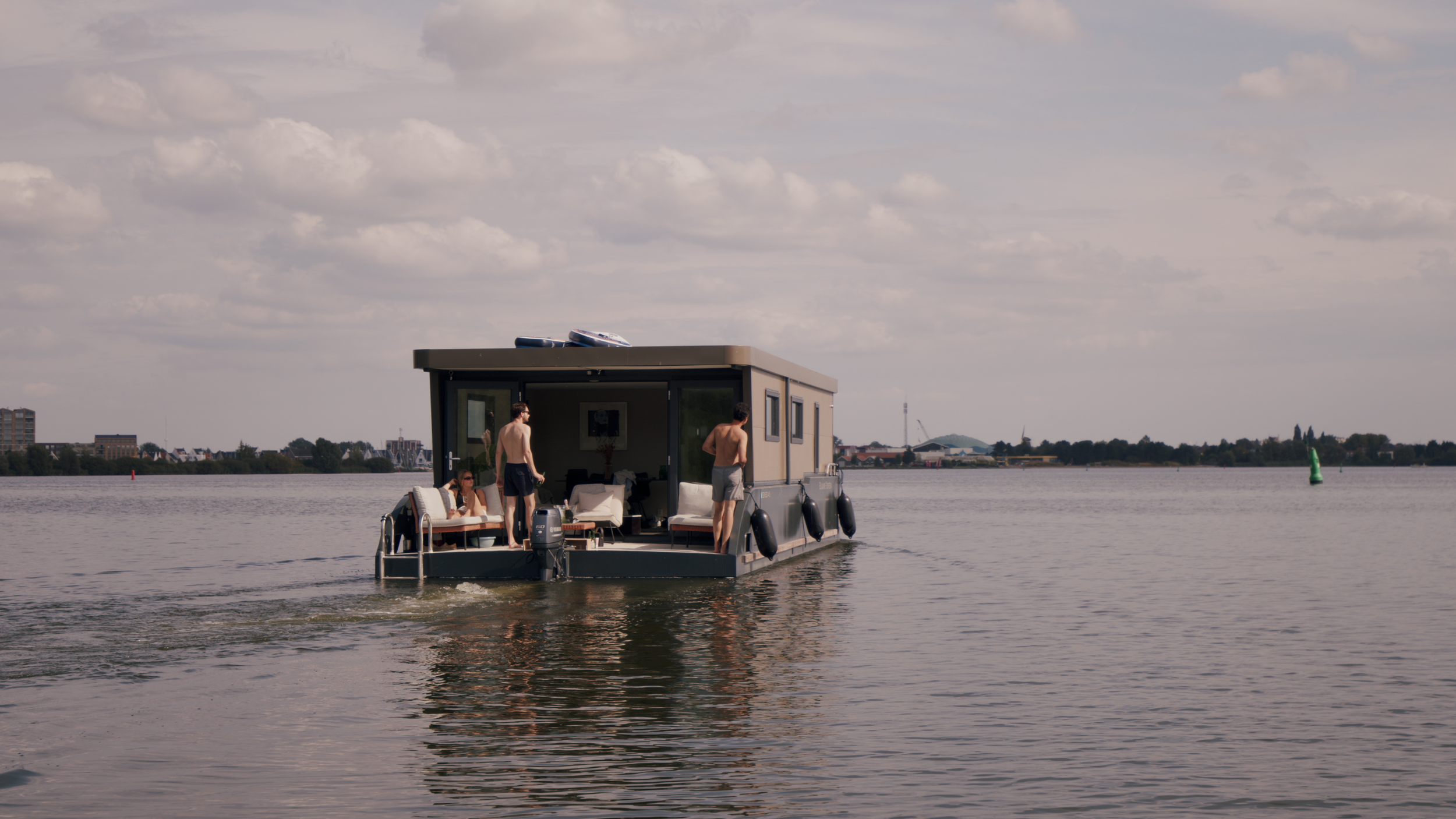 Houseboat with people on deck, cruising on a calm lake under a cloudy sky, distant buildings on the shoreline.