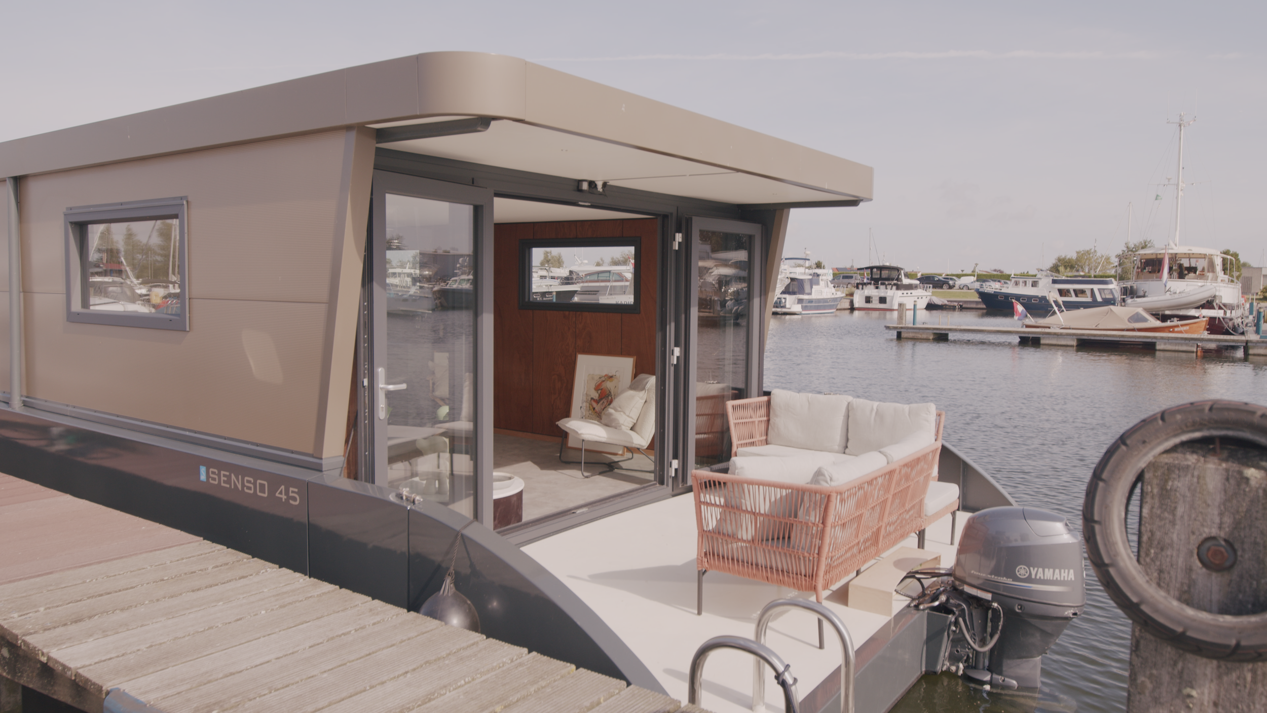 Modern houseboat moored at a marina, with outdoor seating and open sliding doors.