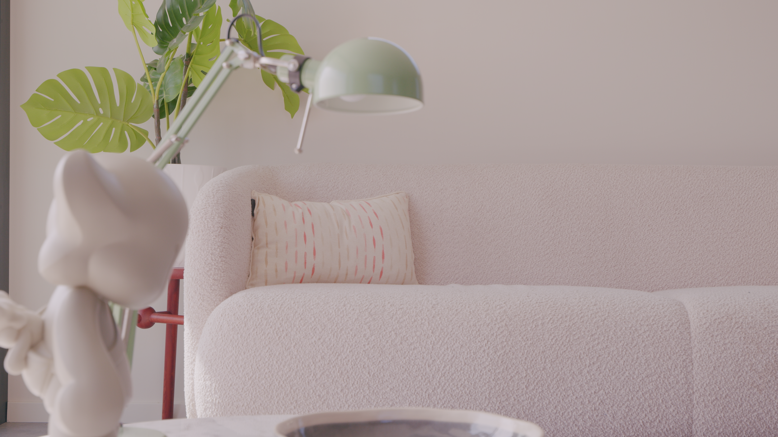 Modern living room with a plush white sofa, decorative pillow, potted plant, green desk lamp, and a small figurine on the table.