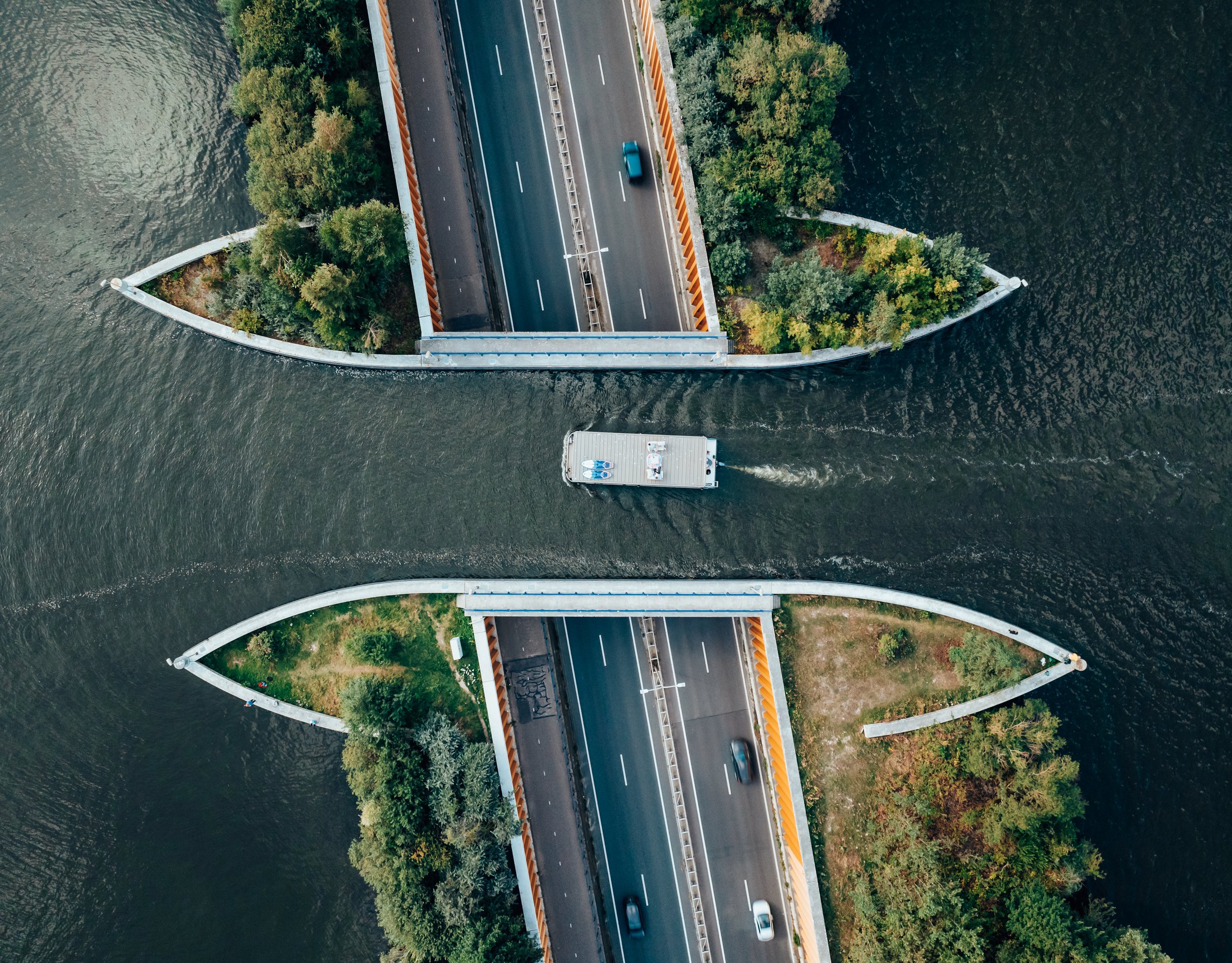 Aerial view of a houseboat navigating a canal passing under two parallel highway bridges, surrounded by lush greenery.
