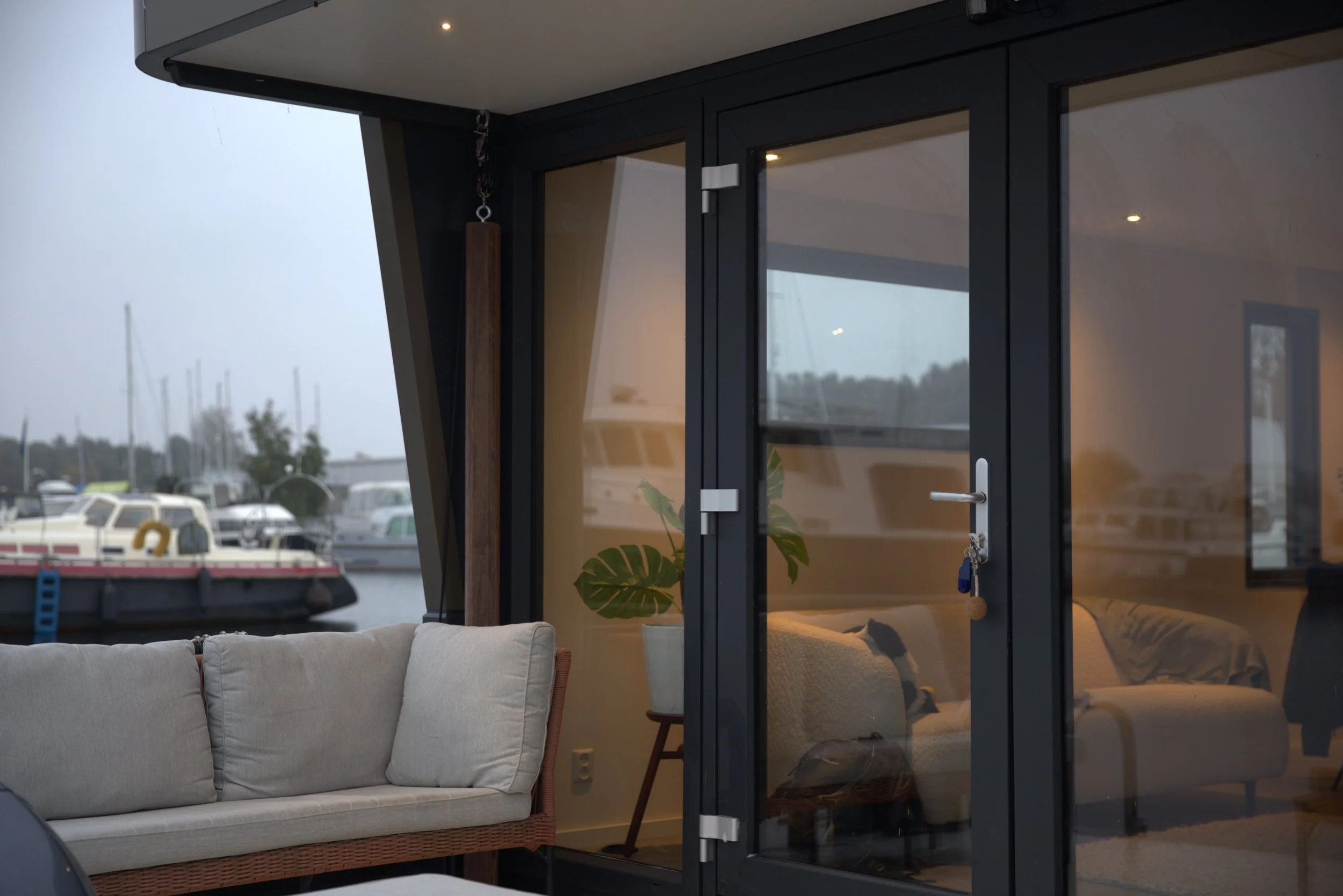 Modern houseboat interior with glass doors, a sofa, and potted plant, docked at a marina with boats visible outside.