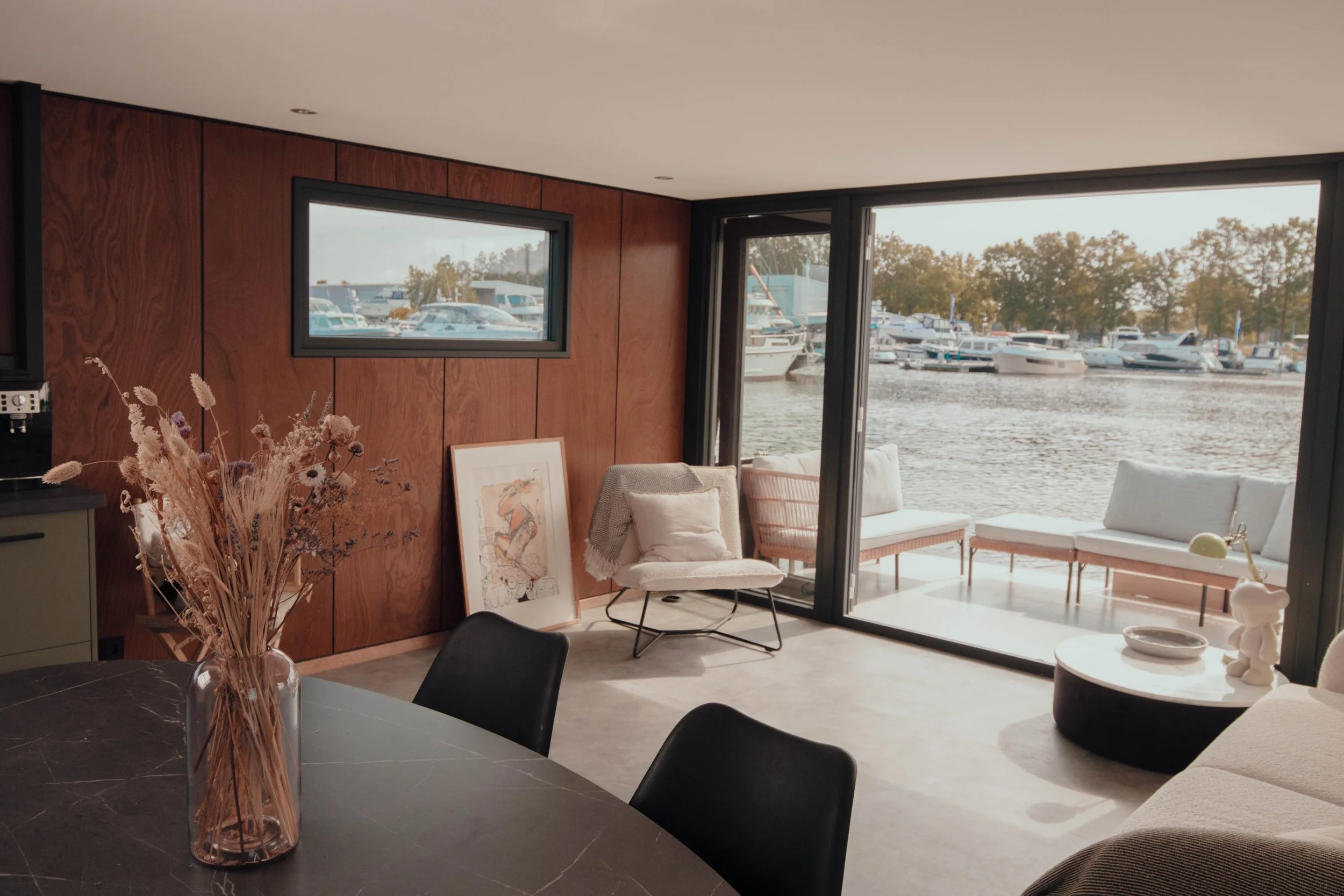 Interior of a modern living room with a view of a marina through large windows, featuring wooden paneling, a round table with a vase of dried flowers, framed art, and contemporary furniture.