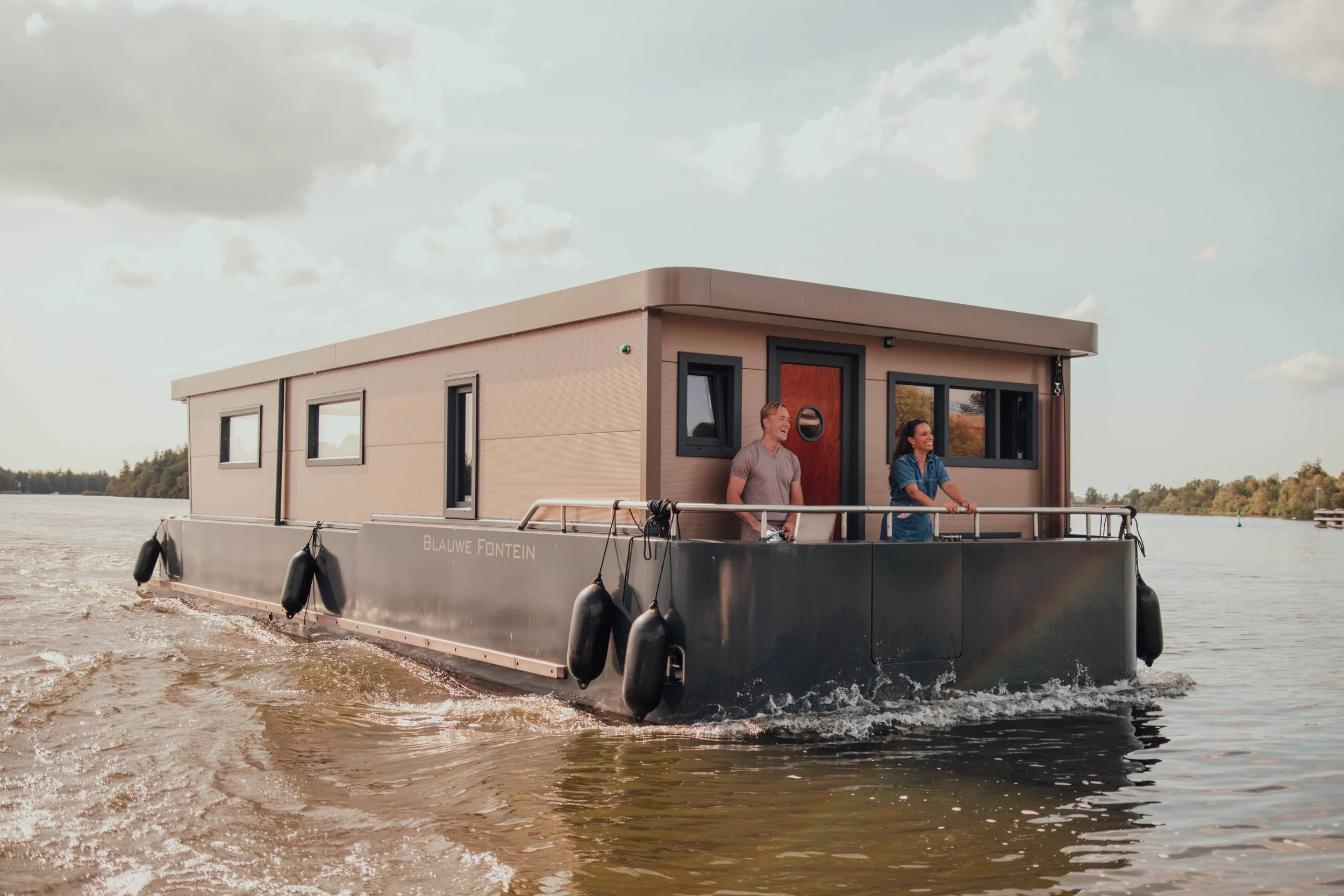 Modern houseboat with people standing on deck sailing on a lake under a cloudy sky.