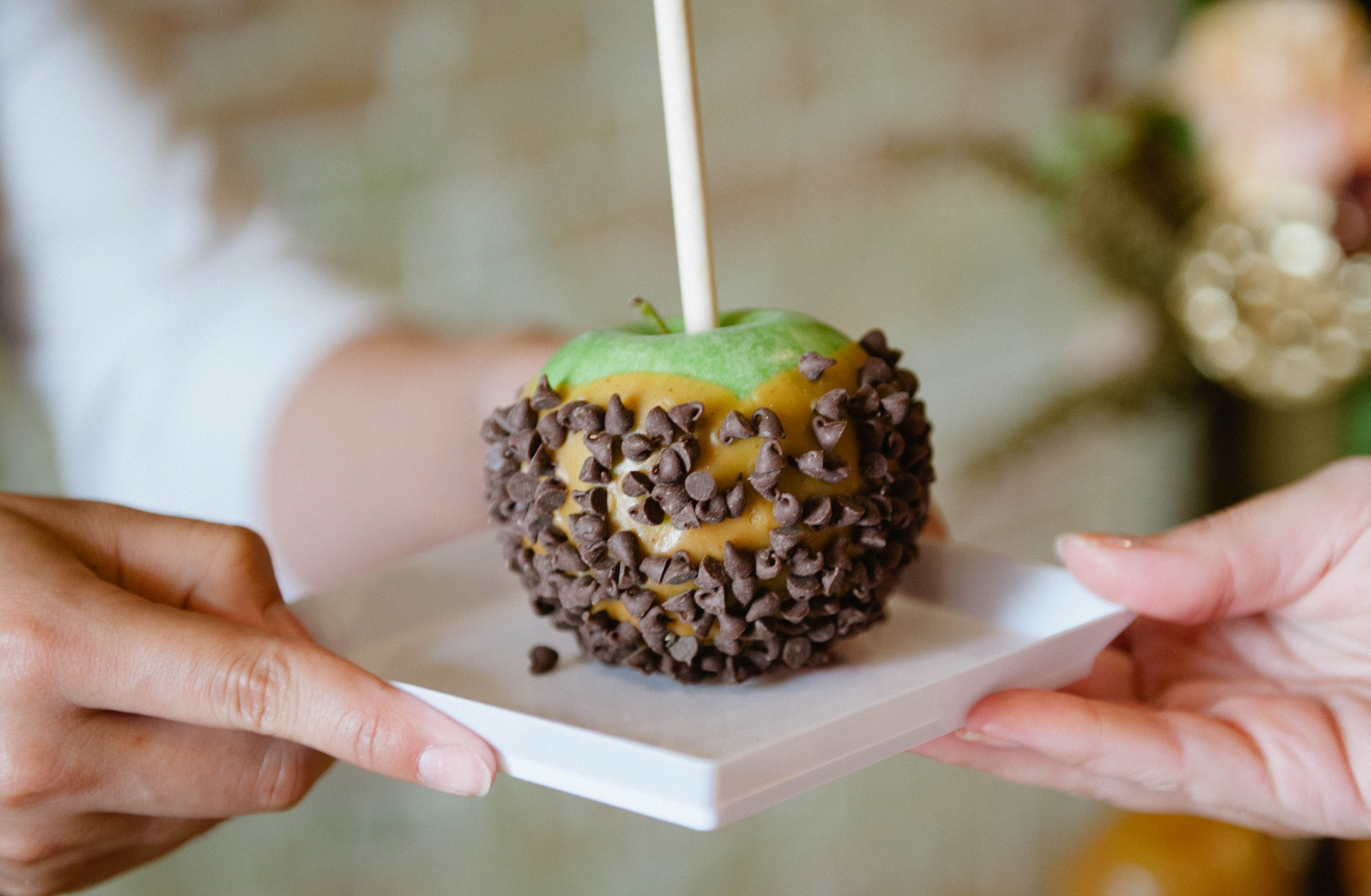 Hands passing a green caramel apple covered in chocolate chips on a small white plate.