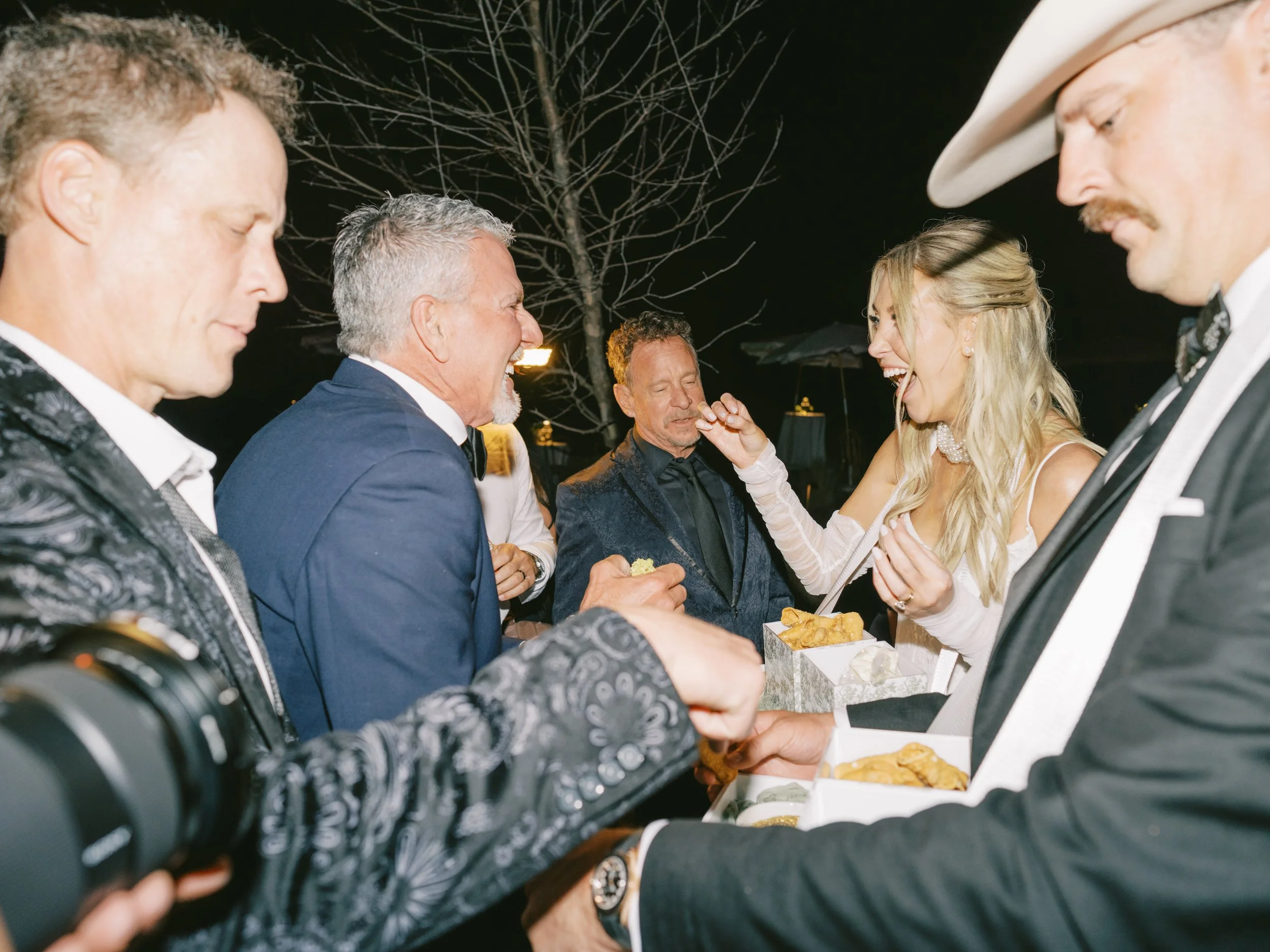 Bride shares a cannoli with her father during an outdoor evening wedding reception while the groom serves fresh-filled cannoli from a custom Roaming Cannoli tray. The Grand Lady, Manor TX