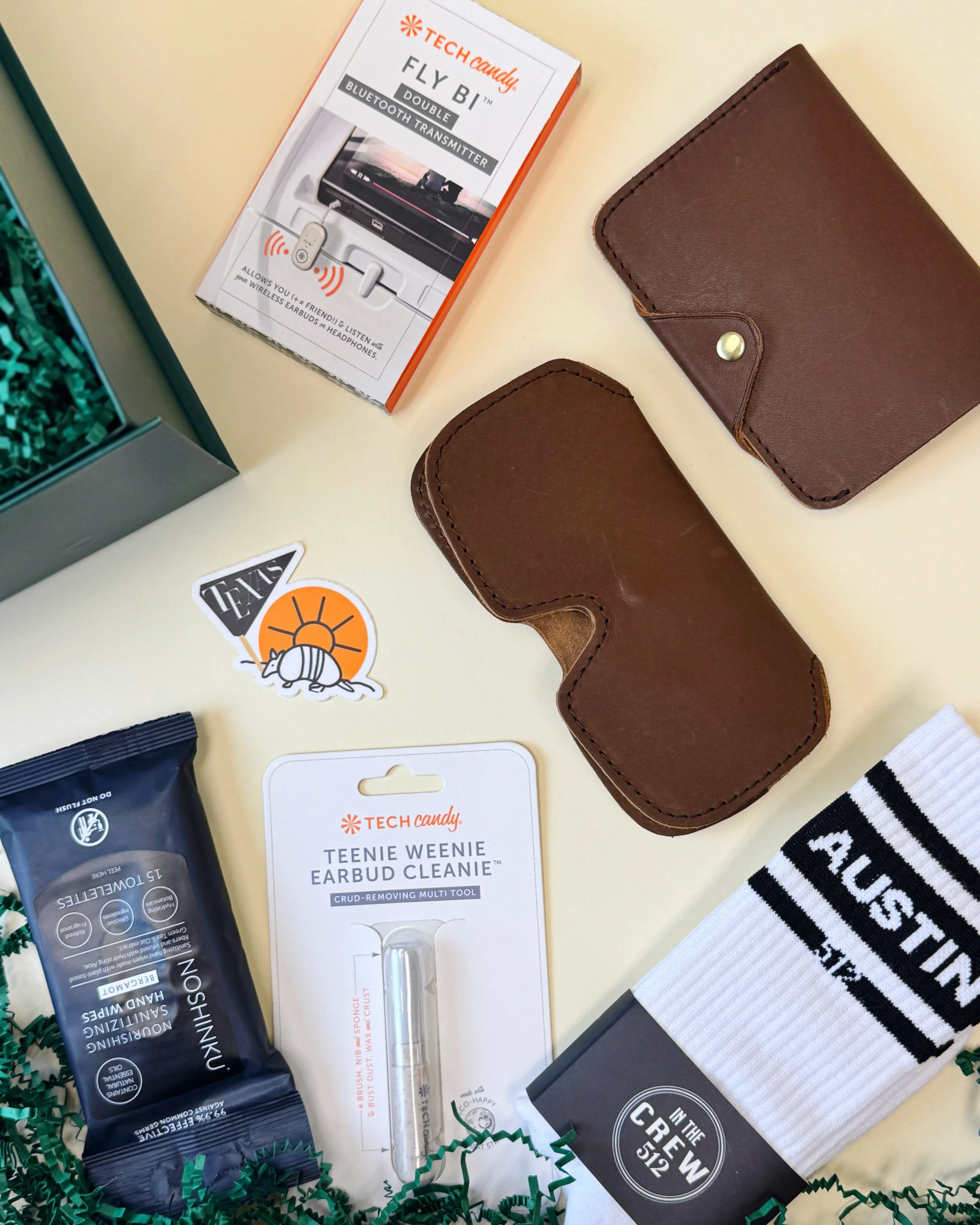 Travel-themed gift box featuring Austin socks, locally made leather sunglasses case and passport case, Bluetooth transmitter, tech cleaning tools, and sanitizing wipes arranged in a green gift box.