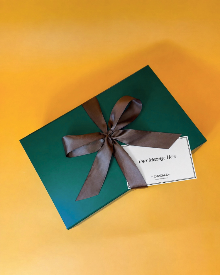 Green gift box with brown satin ribbon on a golden yellow background with a handwritten notecard, part of The Cupcake Bar’s Cozy Autumn Retreat Gift