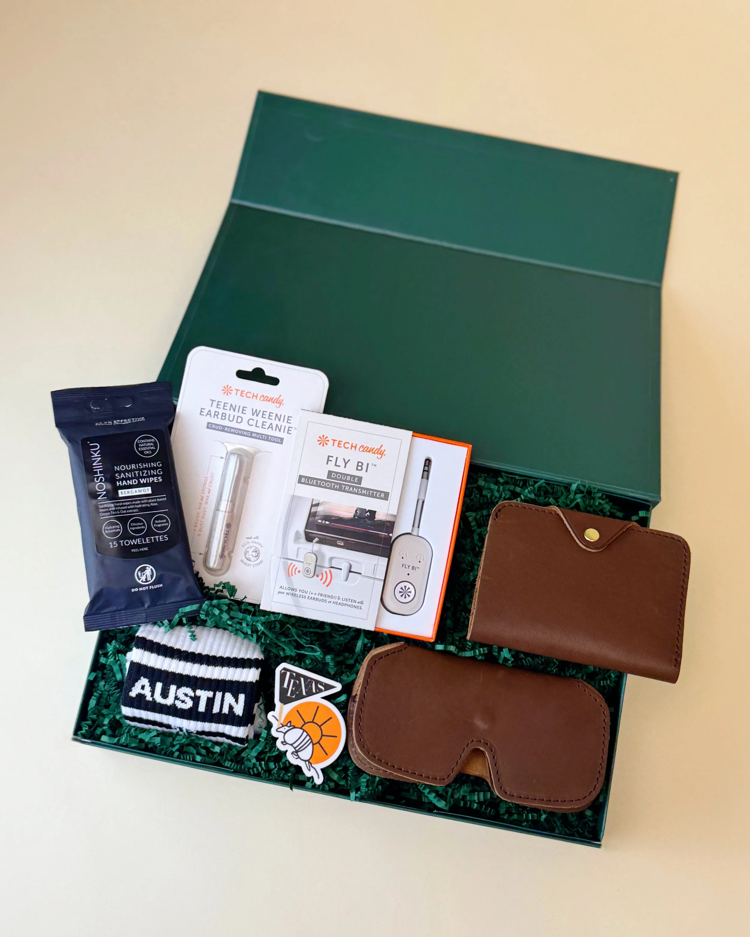 Travel-themed gift box featuring Austin socks, locally made leather sunglasses case and passport case, Bluetooth transmitter, tech cleaning tools, and sanitizing wipes arranged in a green gift box.