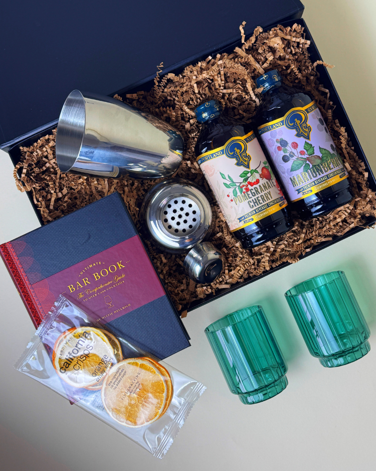 Merry Cocktail features a bar cocktail book, two flavored holiday syrups, a cocktail shaker, glasses, and dehydrated oranges.