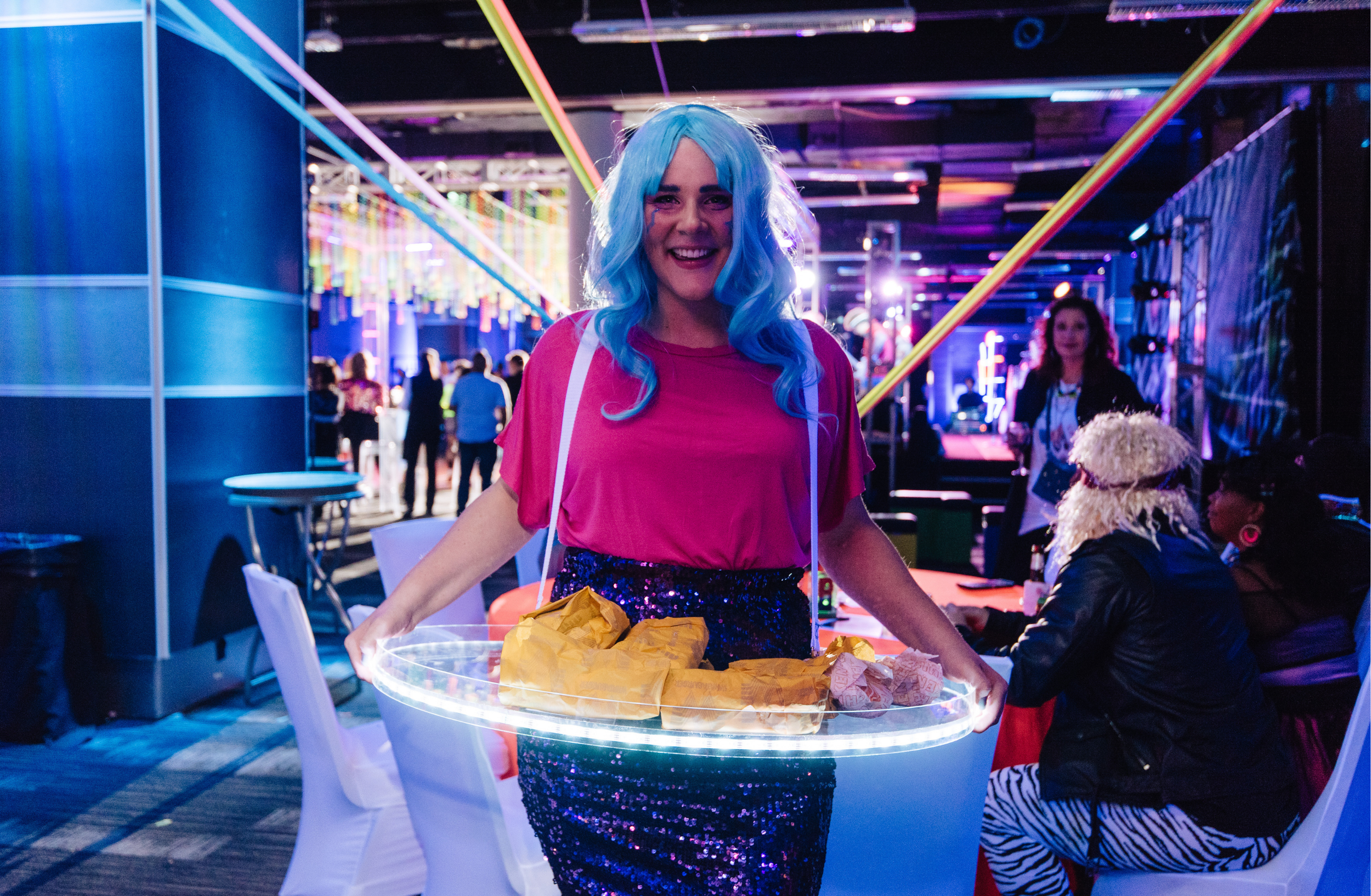 An event attendant wearing a blue wig carries an illuminated roaming tray of bite-sized treats through a dark venue.