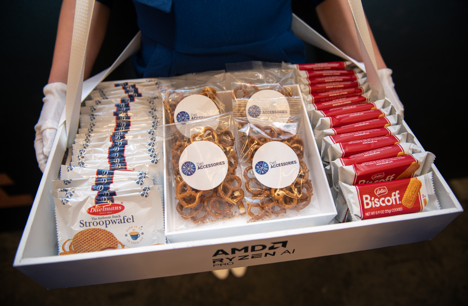 A person holding a branded tray labeled 'AMD Ryzen PRO,' containing individually wrapped stroopwafels, pretzels in plastic bags, and Biscoff cookies.