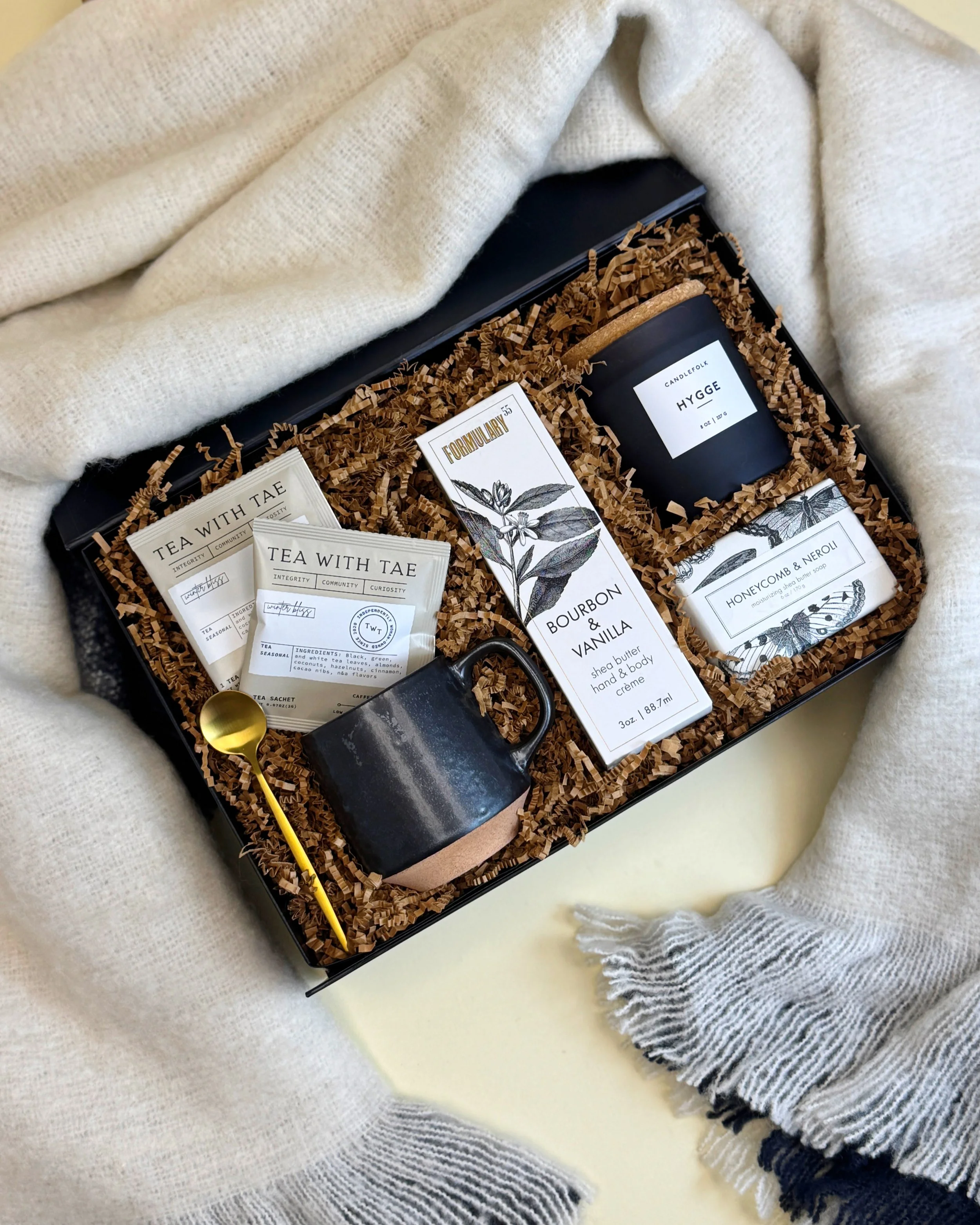 A cozy hygge gift box, including Tea with Tae tea, Formulary 55 soap and lotions, a Hygge candle, soft blanket, and coffee mug and gold-toned spoon