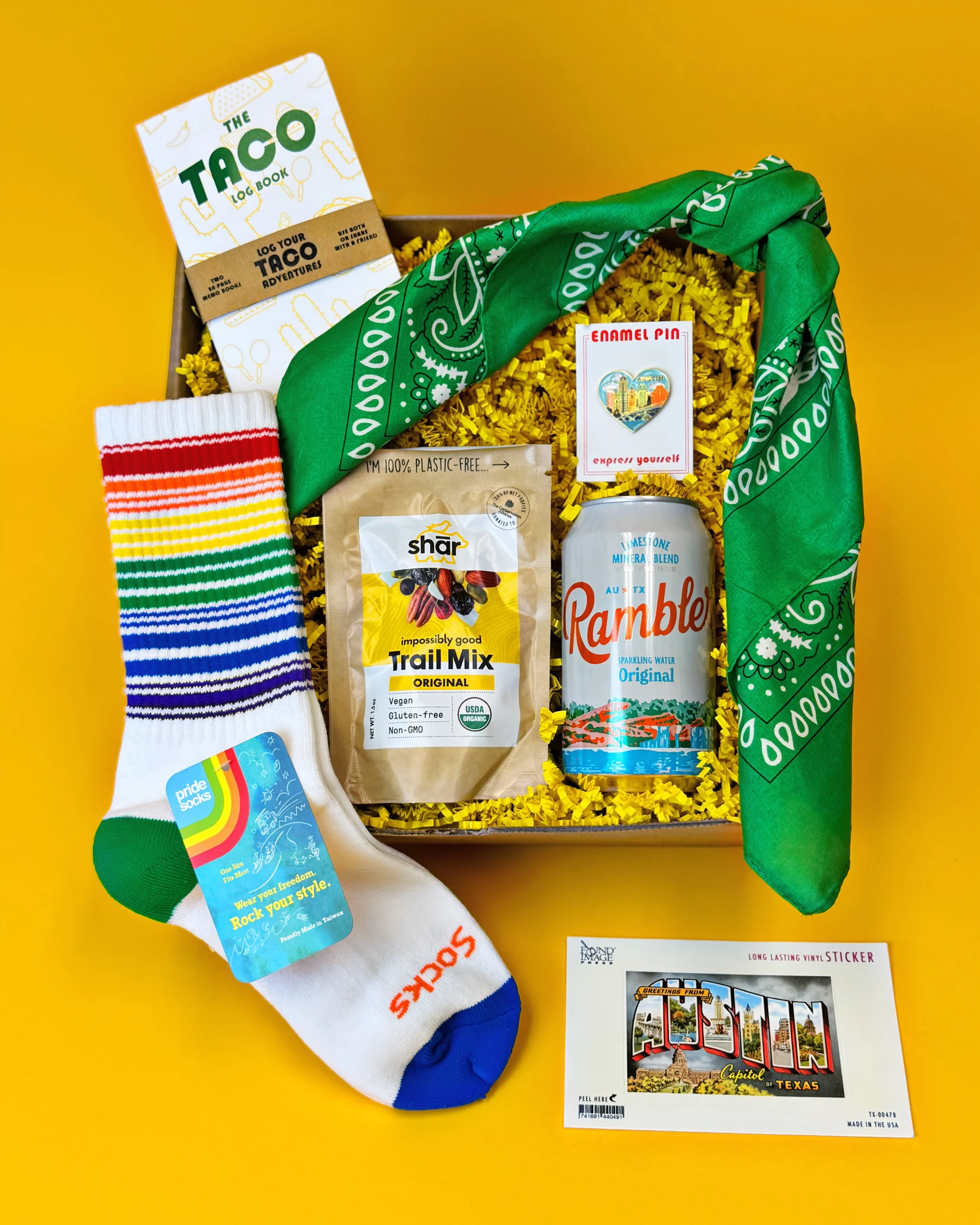 The Austin Greenbelt Gift contains Taco Log Book, Bandana, Rambler Water, Shar Trail Mix, Austin Heart Enamel Pin, Pride Socks Rainbow Crew Socks, and iconic Austin mural vinyl sticker