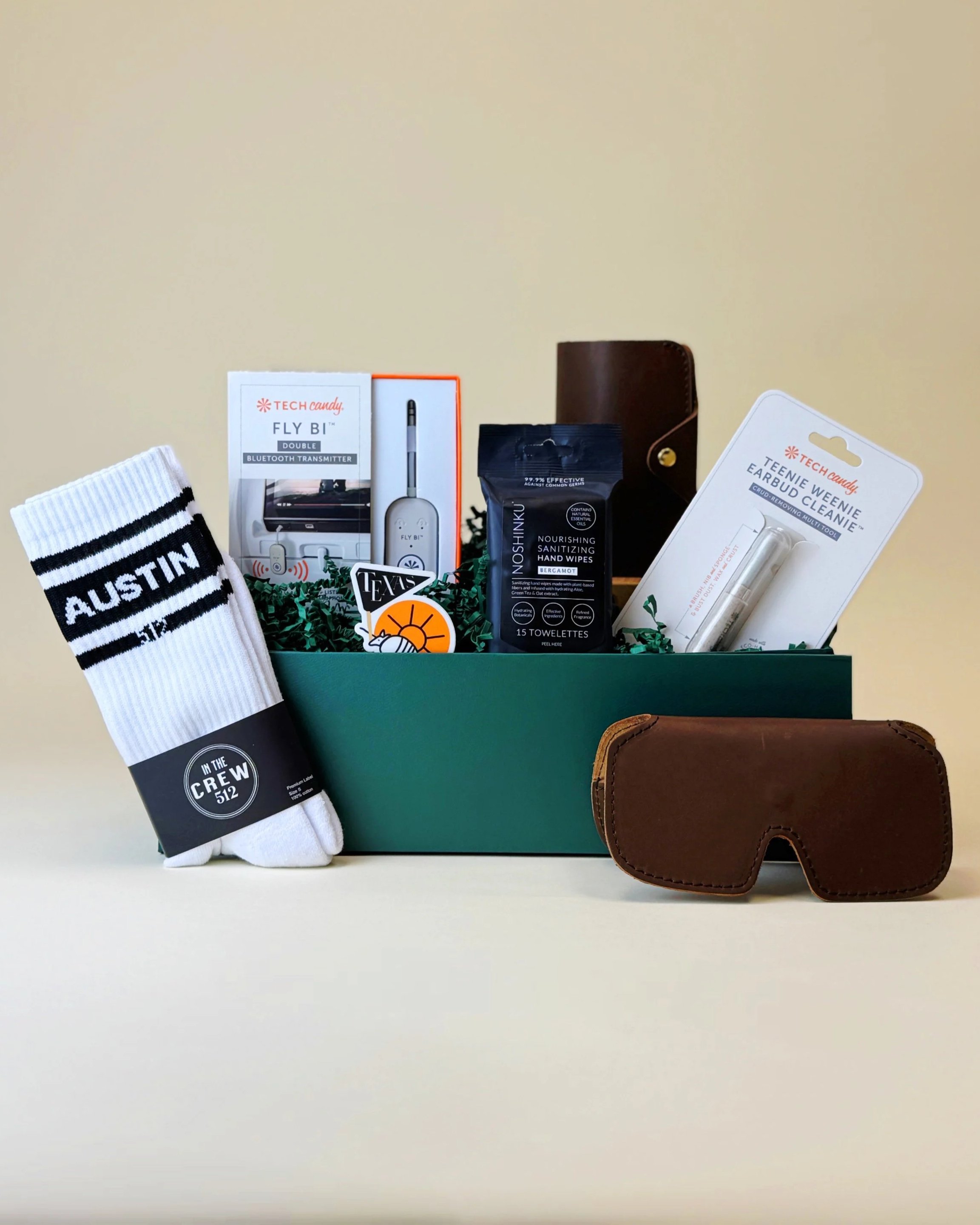Travel-themed gift box featuring Austin socks, locally made leather sunglasses case and passport case, Bluetooth transmitter, tech cleaning tools, and sanitizing wipes arranged in a green gift box.