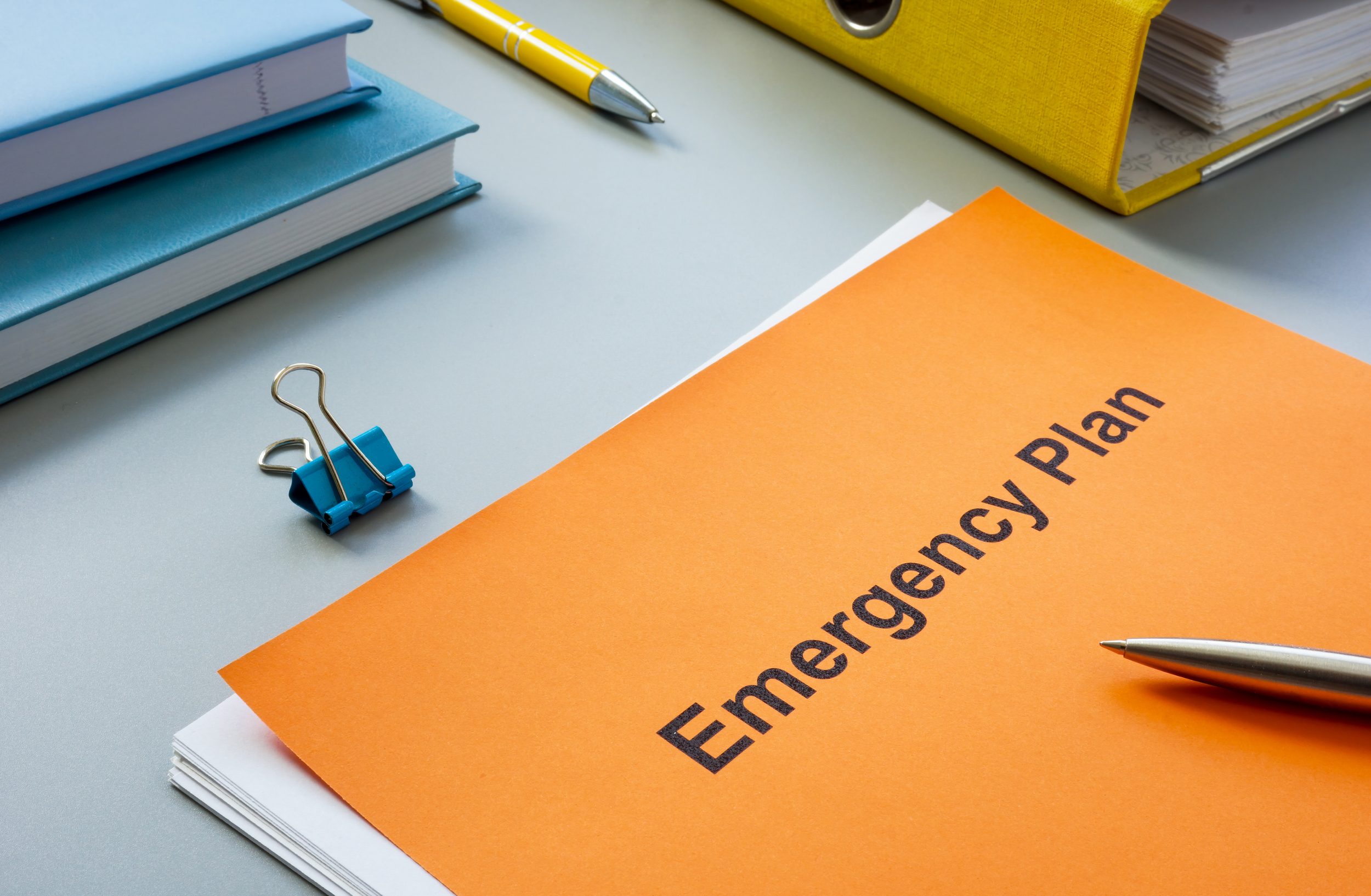 Orange folder labeled Emergency Plan on a desk with notebooks, pens, and office supplies.