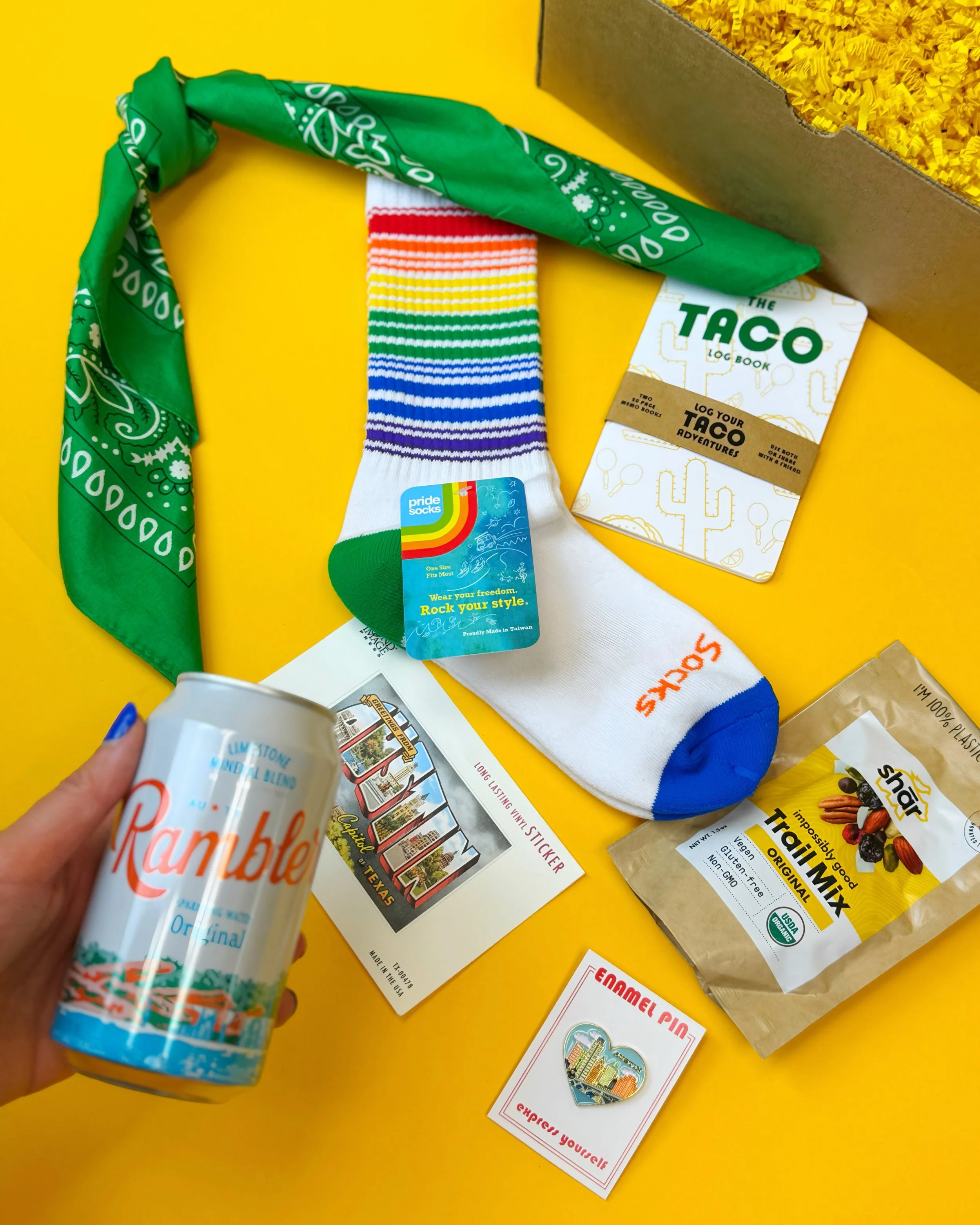 The Austin Greenbelt Gift contains Taco Log Book, Bandana, Rambler Water, Shar Trail Mix, Austin Heart Enamel Pin, Pride Socks Rainbow Crew Socks, and iconic Austin mural vinyl sticker