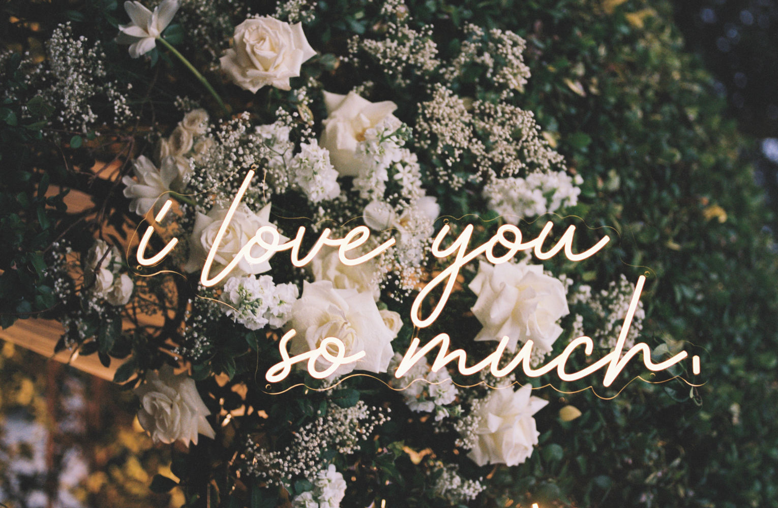 A neon sign with "I love you so much" written in script, a nod to the iconic Austin mural, surrounded by flowers.