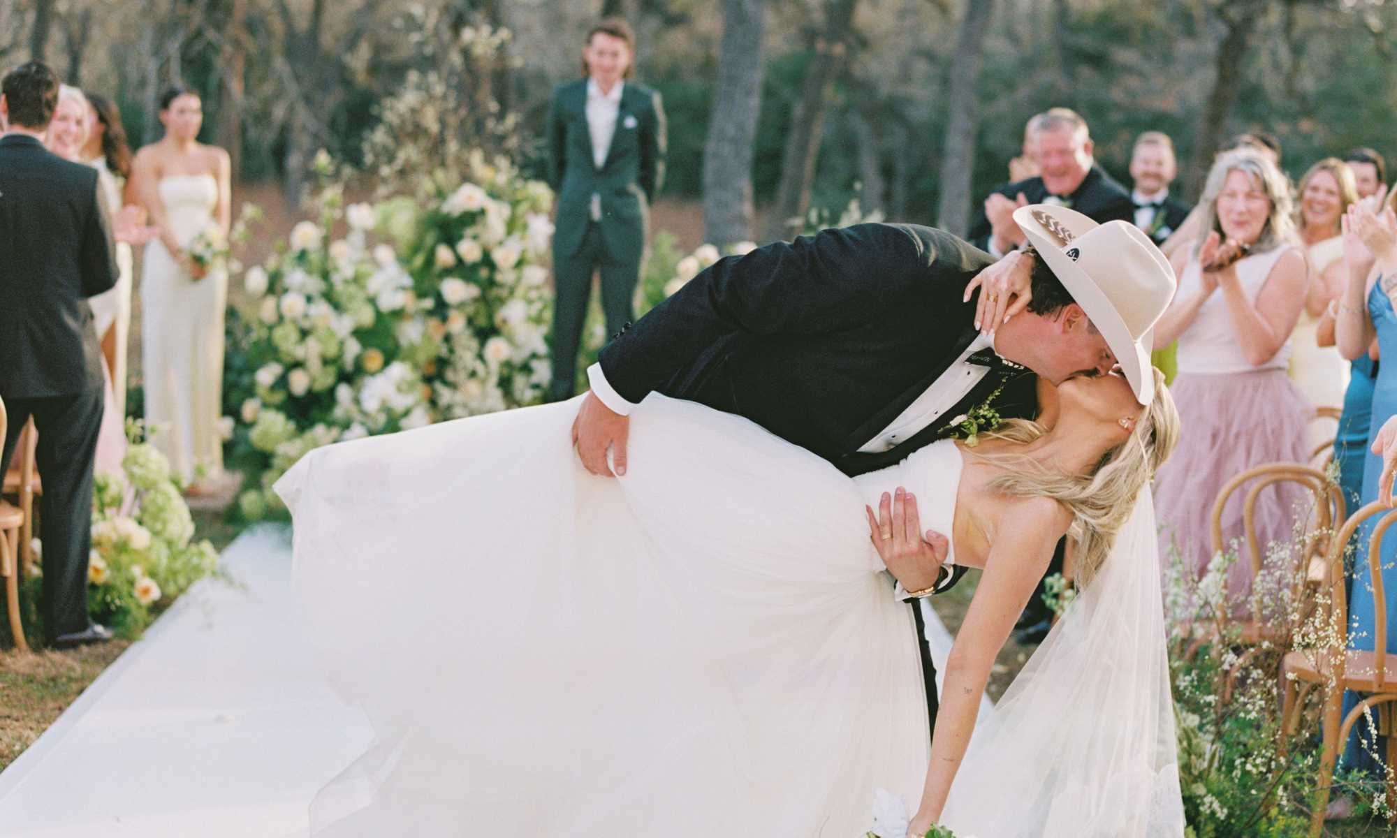 Bride and groom share a kiss during their outdoor wedding ceremony as guests applaud and smile around them.