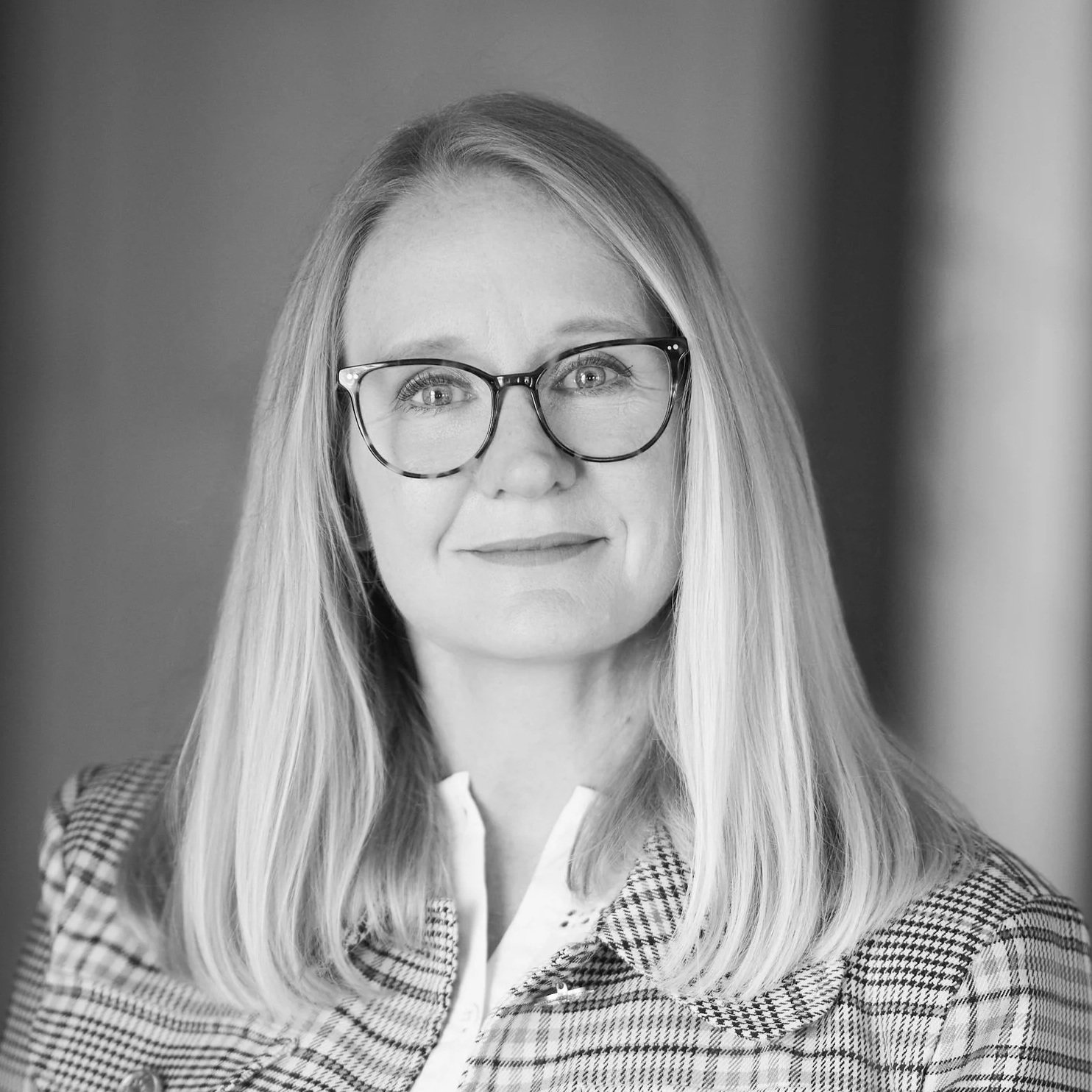 Black and white portrait of a woman with long, straight hair wearing glasses, a checkered blazer, and a white shirt, looking slightly to the side with a subtle smile.