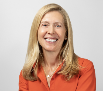 A woman with long blonde hair, smiling, wearing an orange blazer and a necklace, against a light gray background.