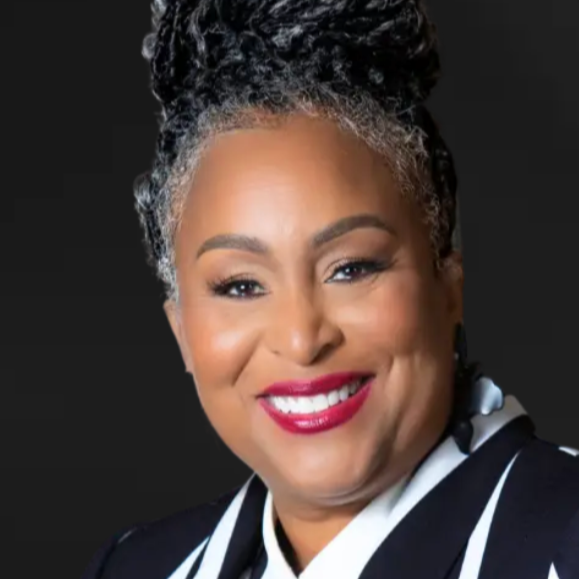 Close-up of a smiling African American woman with curly gray hair in an updo, wearing a black and white striped jacket against a dark background.