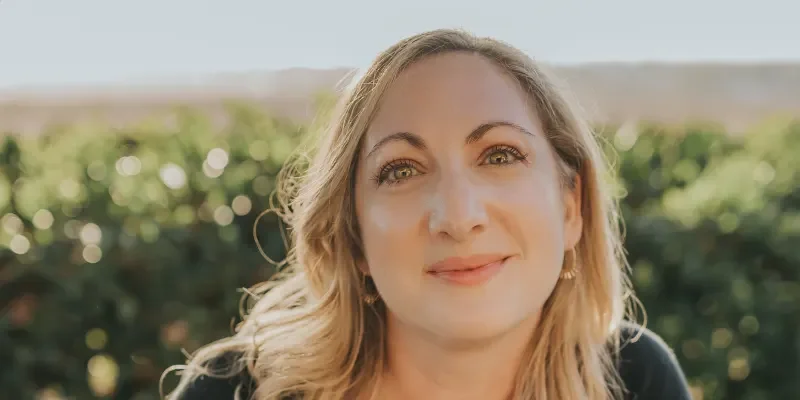 A woman with blonde wavy hair smiling outdoors with a blurred green landscape in the background.