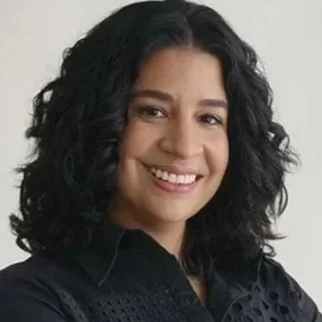 Smiling woman with curly black hair, wearing a dark top, posed against a neutral background.