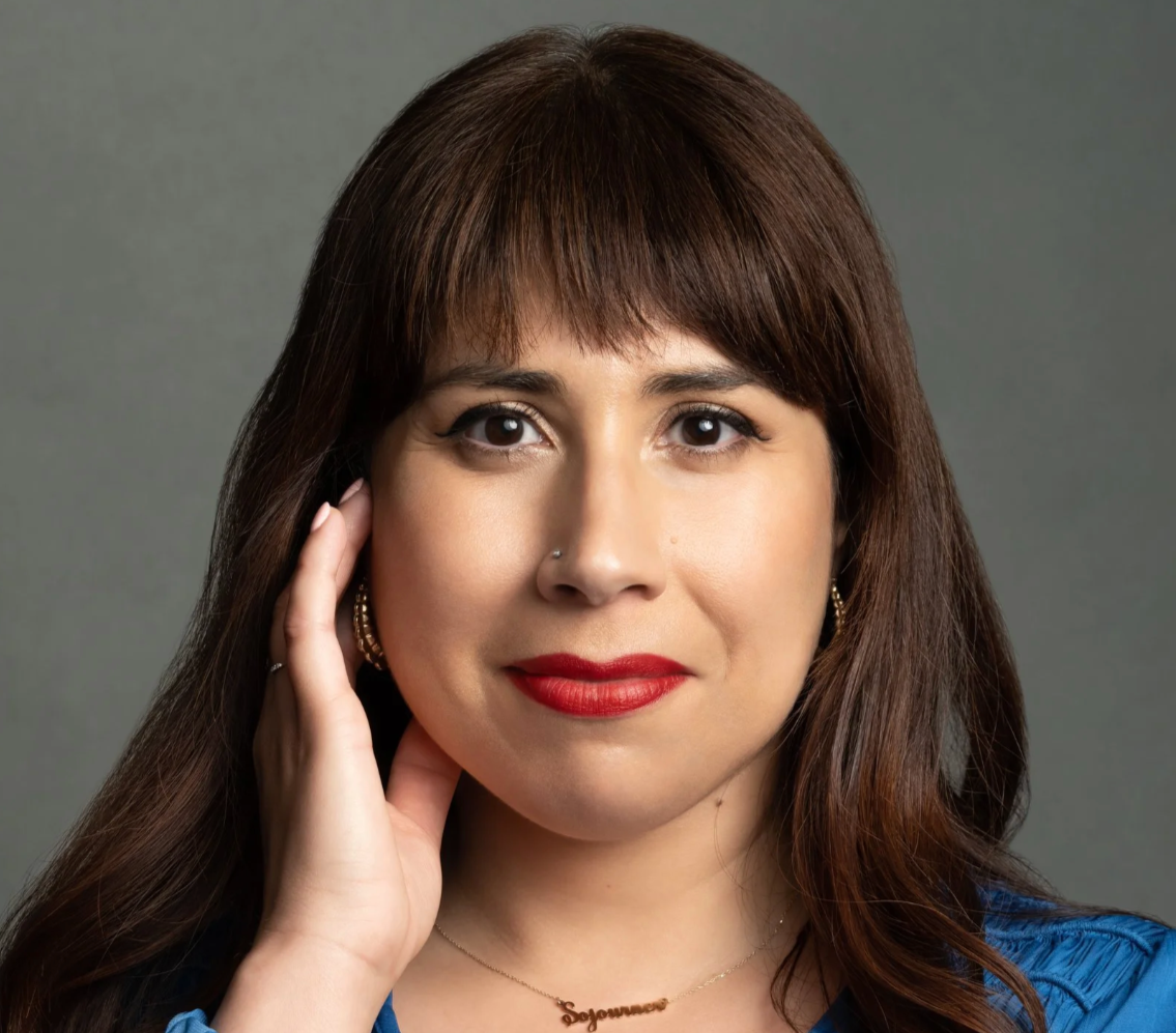 Portrait of a woman with long brown hair, red lipstick, wearing a blue top, gold earrings, a nose piercing, and a necklace that says 'Soyunore', touching her face with her right hand, against a gray background.