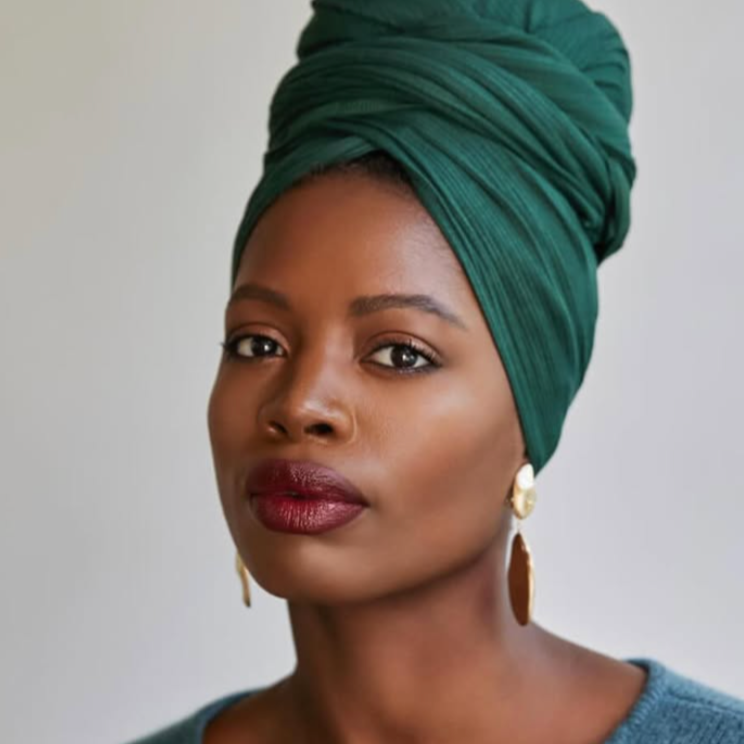Portrait of a Black woman wearing a green headwrap, gold earrings, and a blue top, looking confidently at the camera.