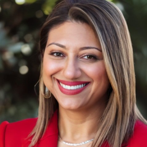 A smiling woman with light brown hair wearing a red blazer outdoors.