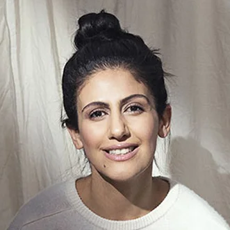 A young woman with dark hair in a bun, smiling, wearing a white sweater, standing in front of a light-colored curtain.