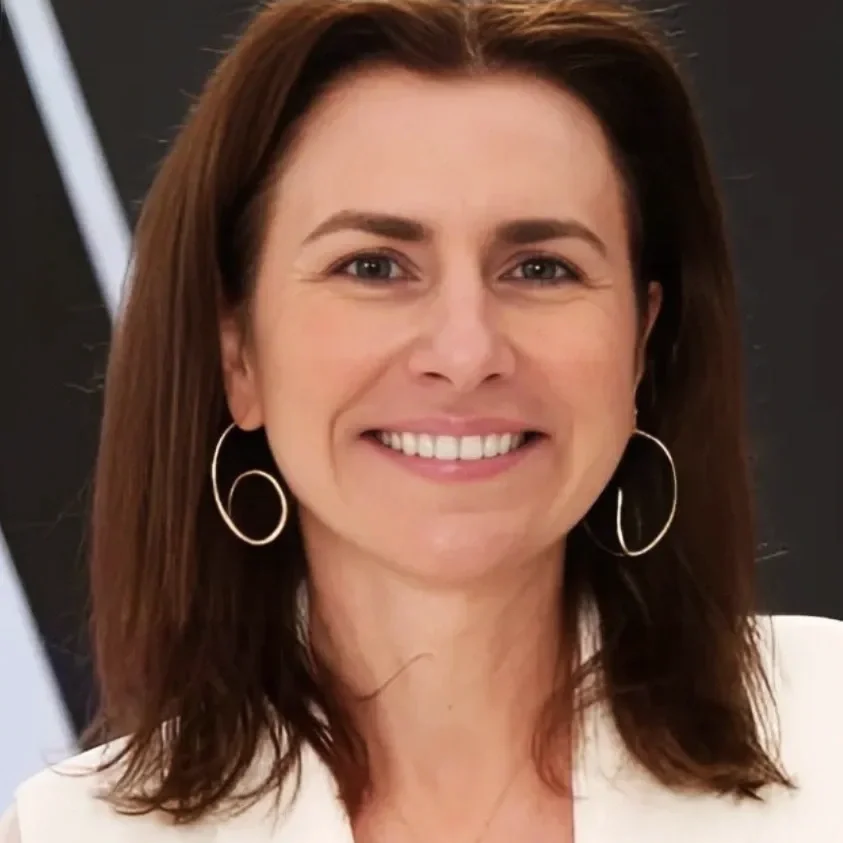 A woman with shoulder-length brown hair smiling, wearing hoop earrings and a white top, standing in front of a black background.