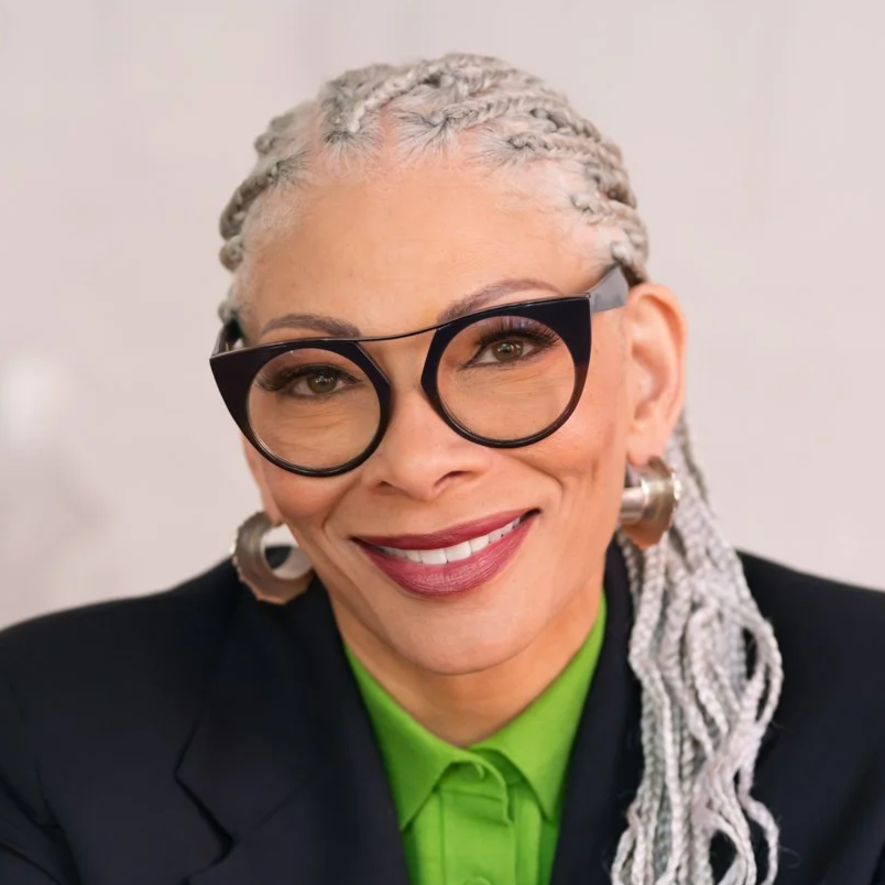 A woman with gray hair styled in braids, wearing black-rimmed glasses, large silver earrings, a green shirt, and a black blazer, smiling at the camera.