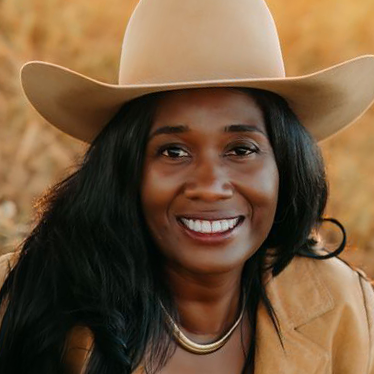 Woman wearing a cowboy hat, smiling, outdoors with a blurred natural background.
