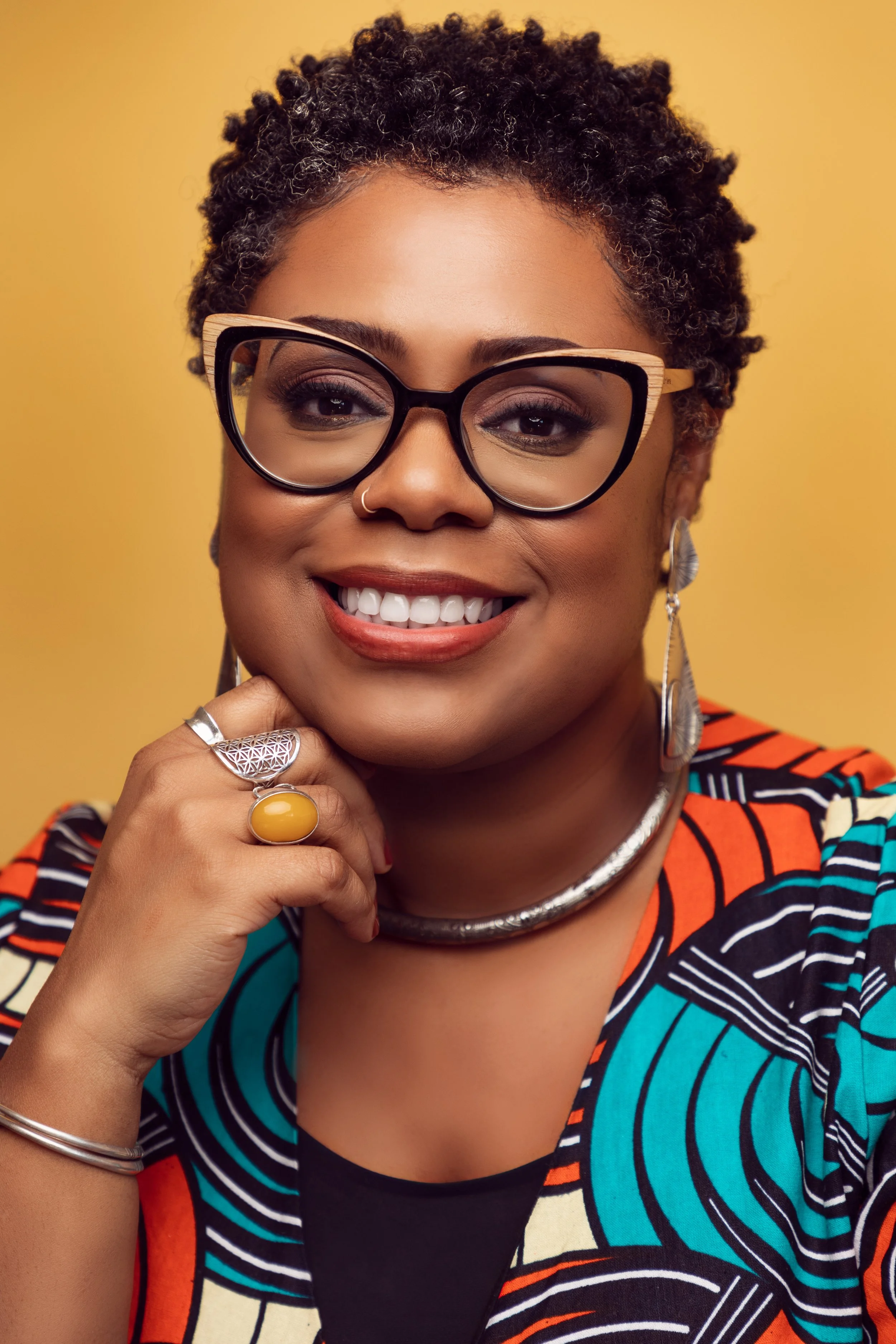 A smiling woman with short, curly hair, wearing large black-framed glasses with a wooden accent on the top, and colorful jewelry including a silver necklace, large earrings, and rings. She is dressed in a vibrant, patterned top and posed against a plain yellow background.