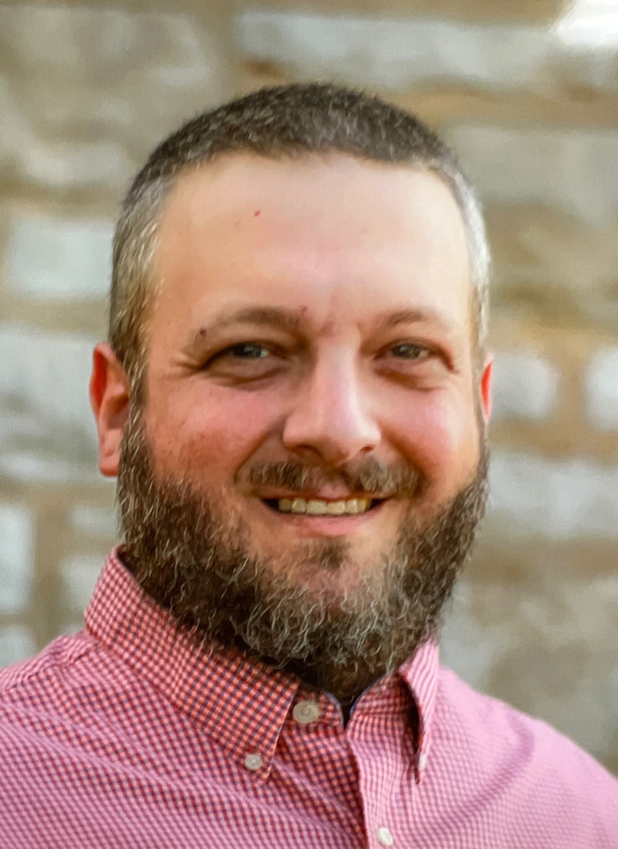 Midlife man with short gray hair and a beard, smiling, wearing a pink and white checkered shirt, in front of a blurred brick wall background.