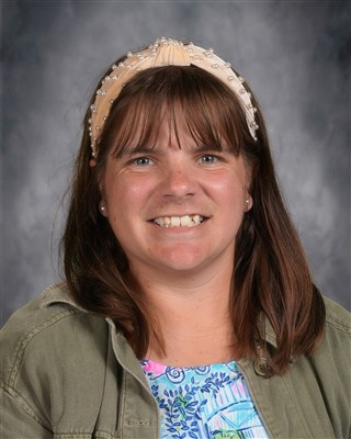 A woman with shoulder-length brown hair wearing a pearl-embellished headband and a green jacket, smiling at the camera.