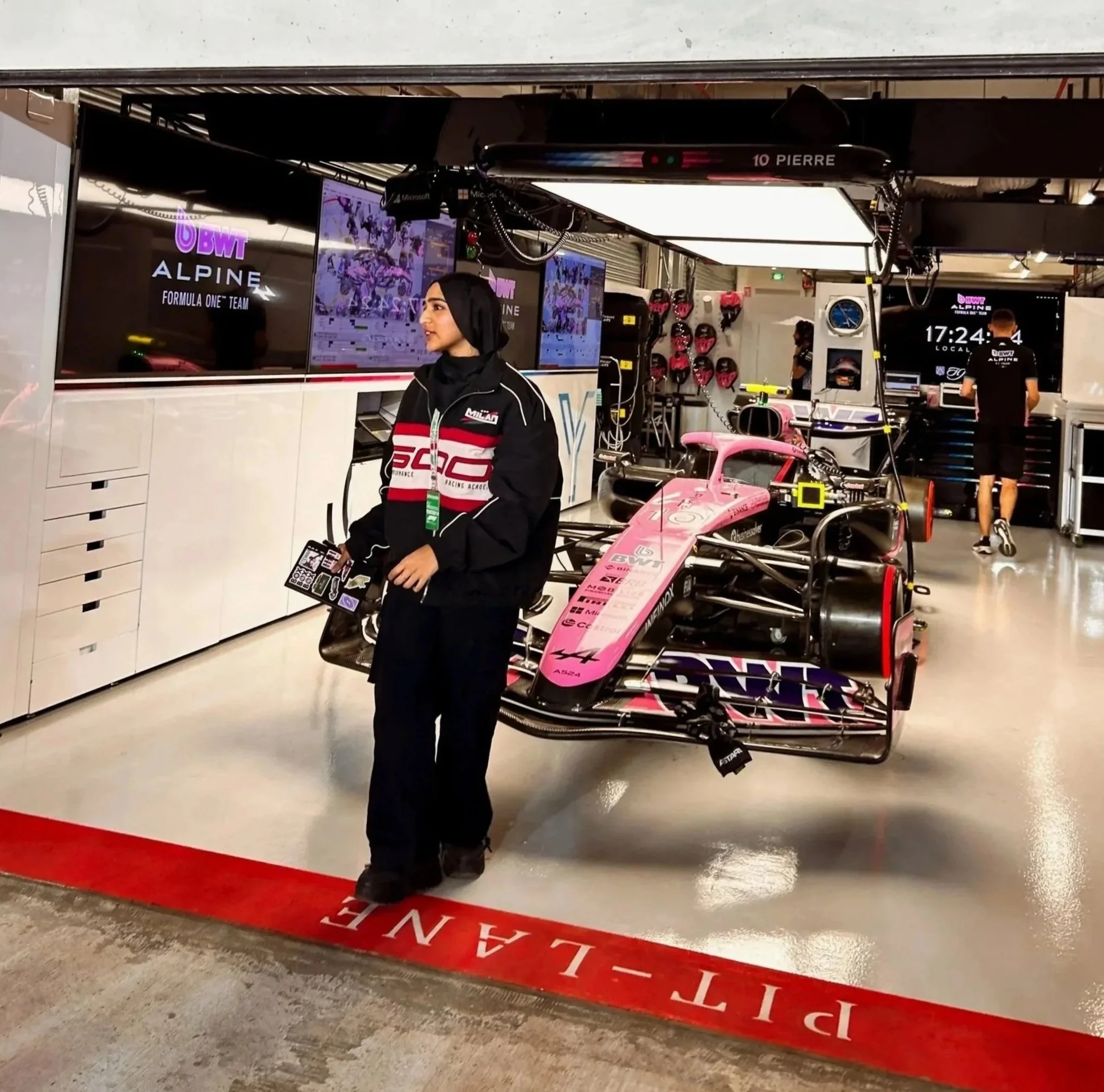 A woman in a black jacket with red and white accents stands in the garage of a race car, with a pink Formula 2 race car behind her. The garage features TV screens, toolboxes, and equipment, with two men working in the background.