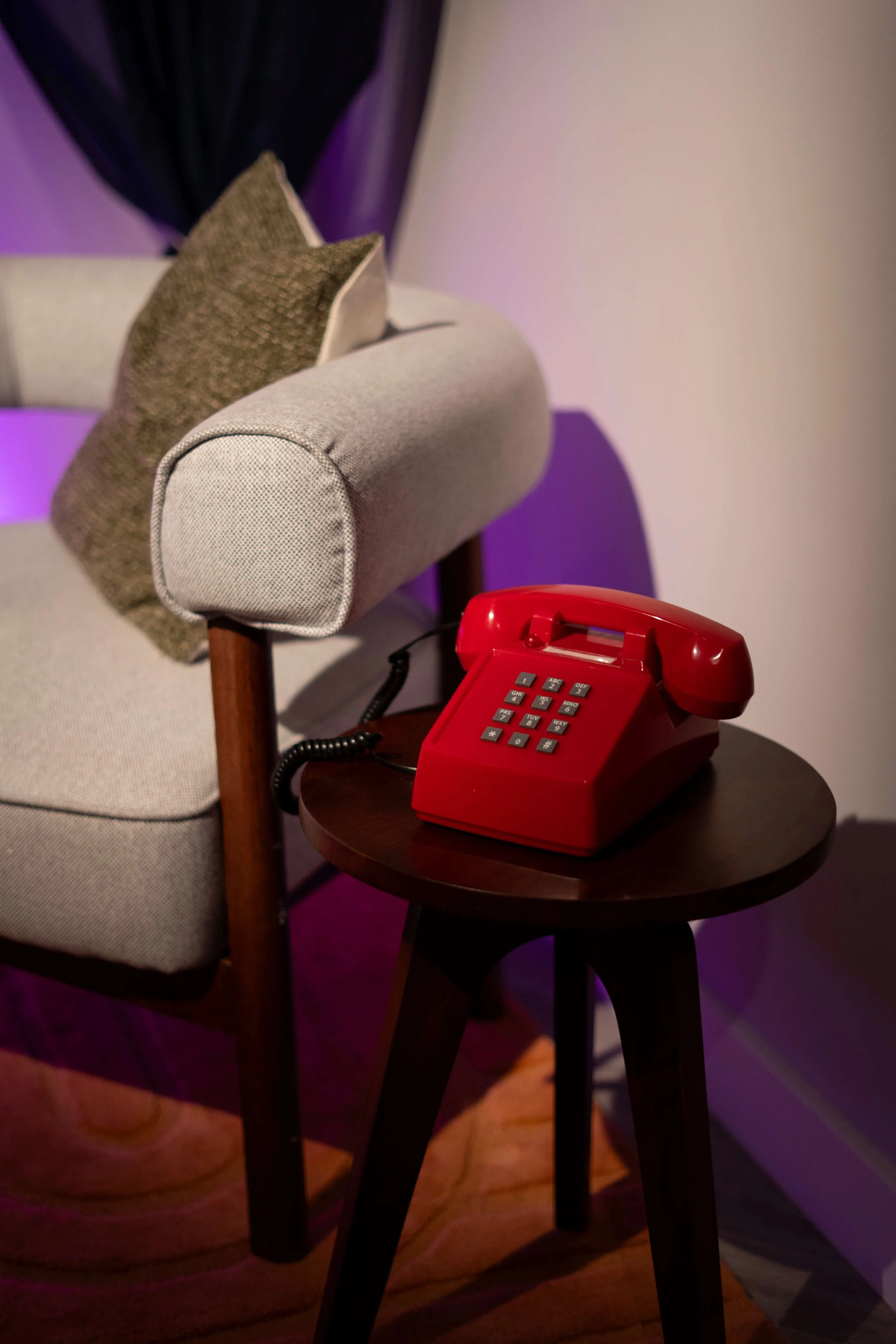 A red vintage rotary telephone on a small round dark wood table next to a beige upholstered armchair with patterned pillows. The scene is lit with purple lighting.