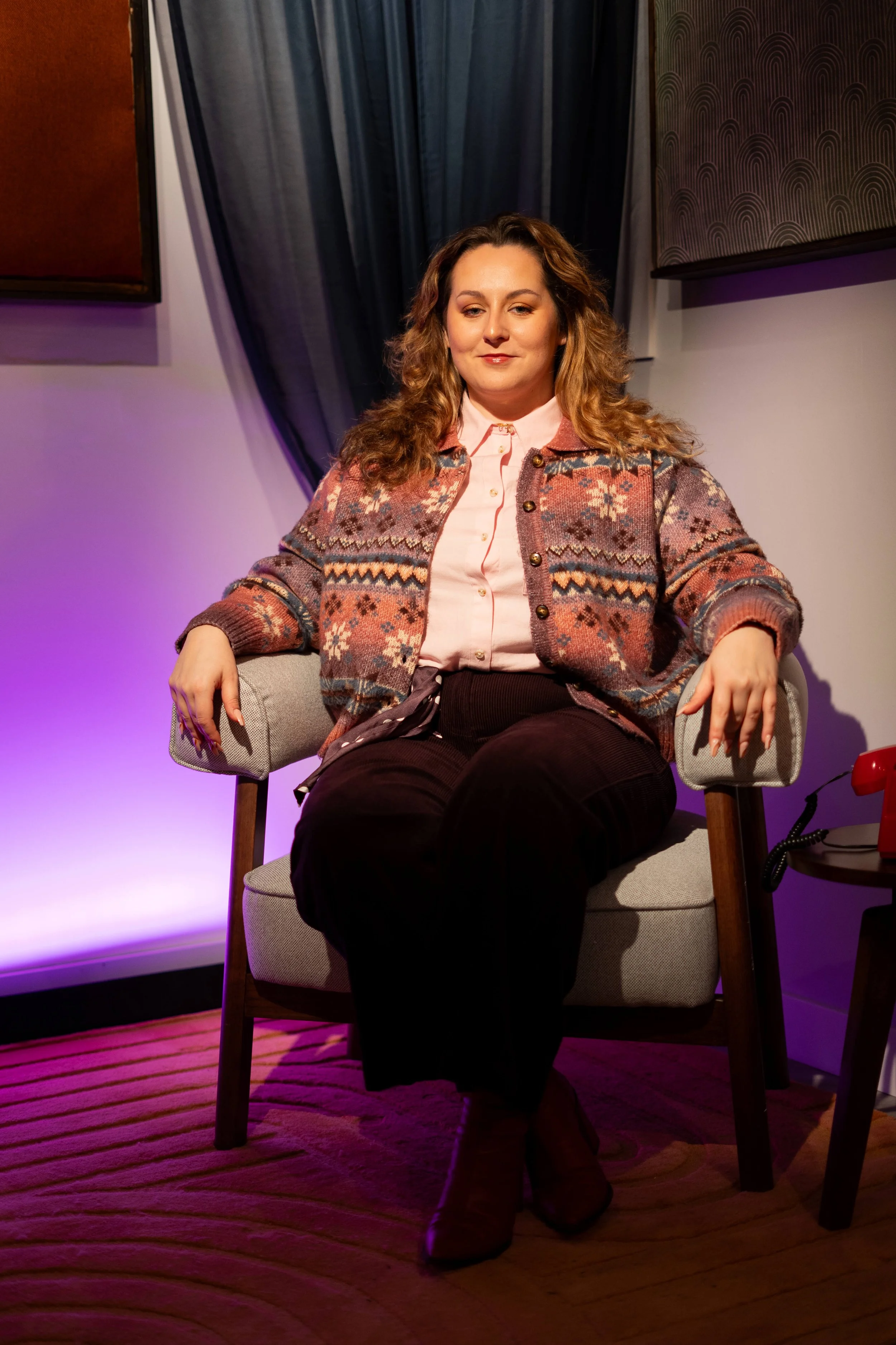 A woman with curly brown hair sitting on a beige armchair in a room with purple and blue lighting. She is wearing a pink shirt and a patterned cardigan, with her legs crossed and hands resting on the armrests.