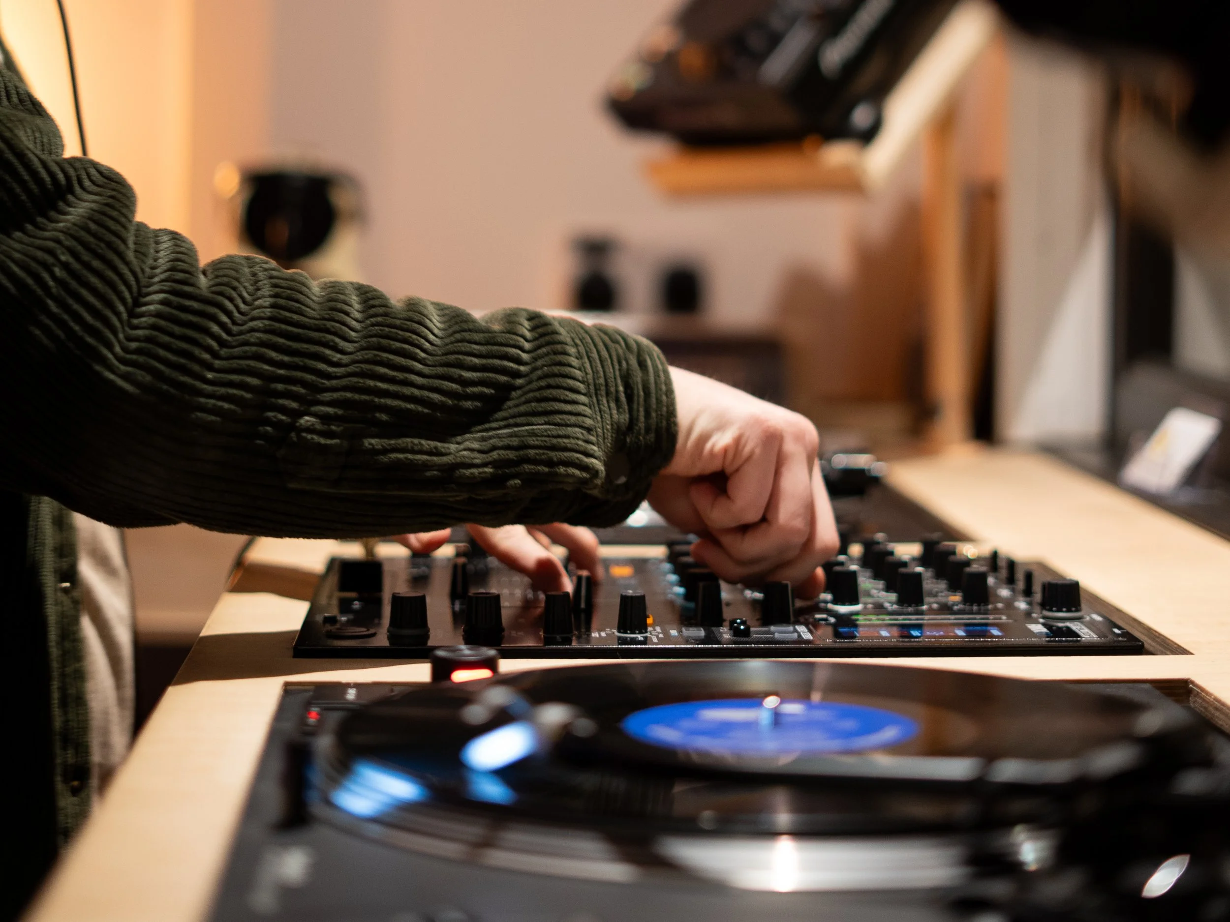 A person wearing a green sweater operating a DJ mixer with vinyl records in the foreground and a blurred background.