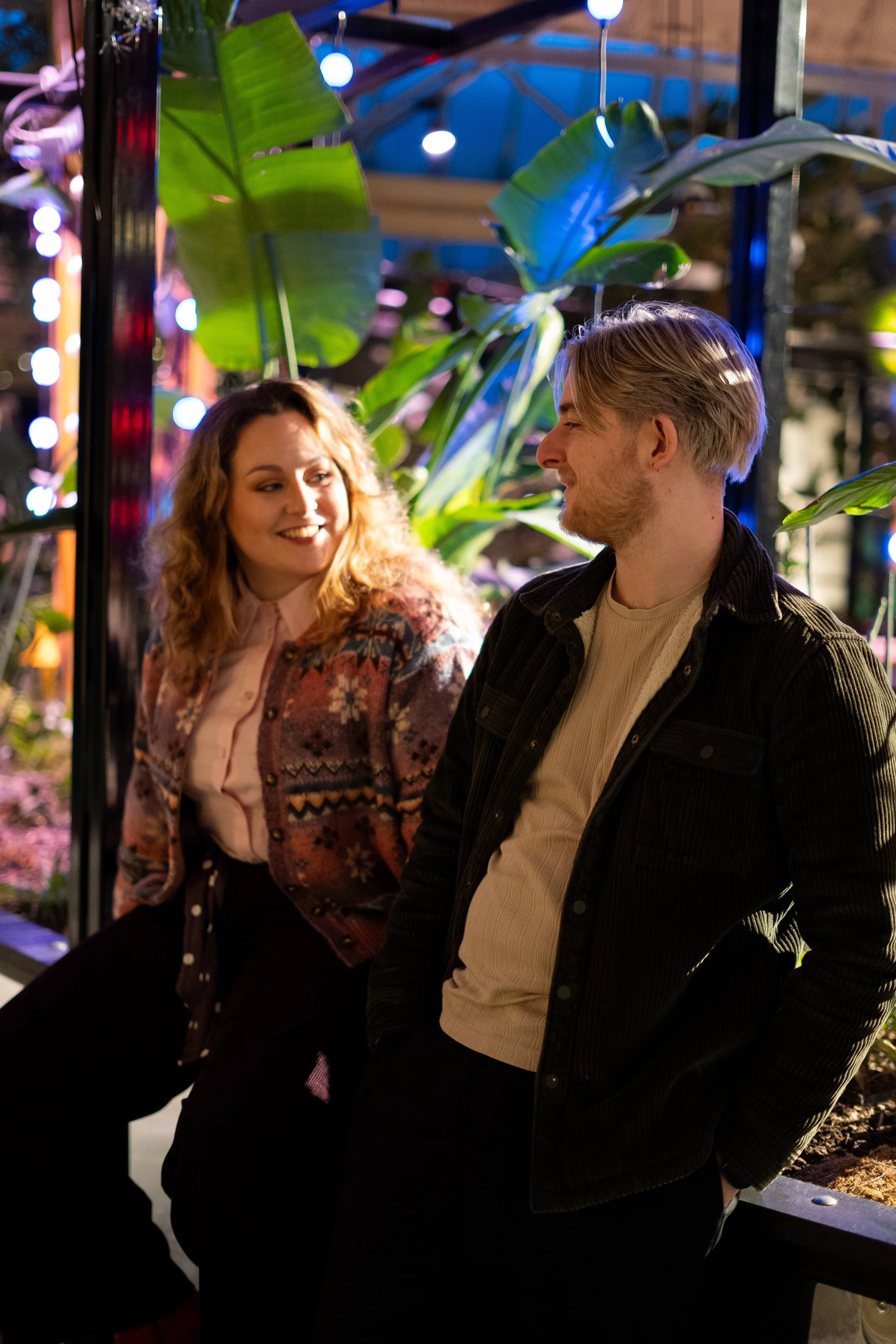 A woman with curly hair smiling at a man with blond hair, both standing under large green leaves at night, with string lights in the background.