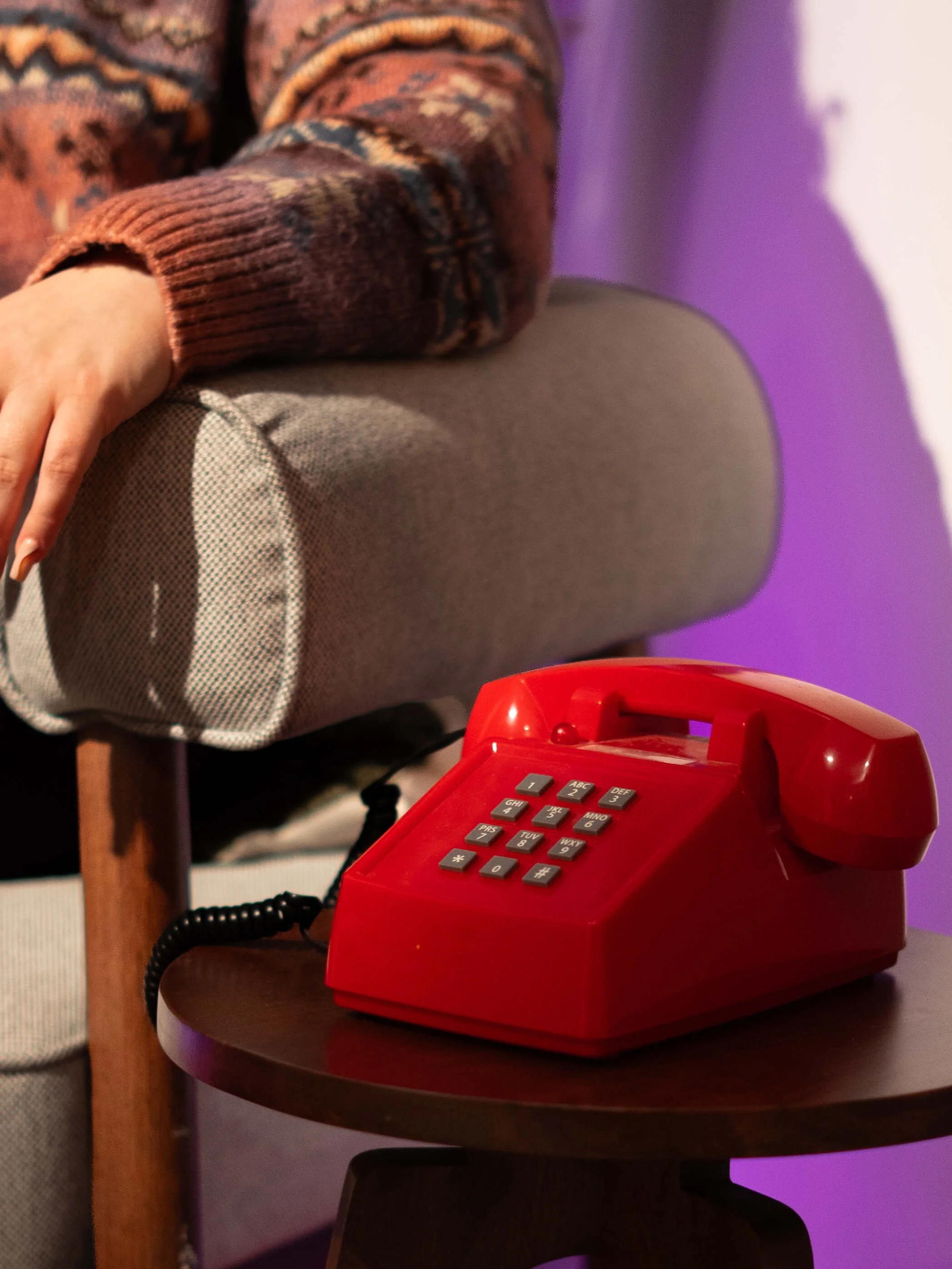 Red vintage rotary telephone on a dark round table, in a room with a person wearing a colorful sweater sitting nearby and purple lighting in the background.