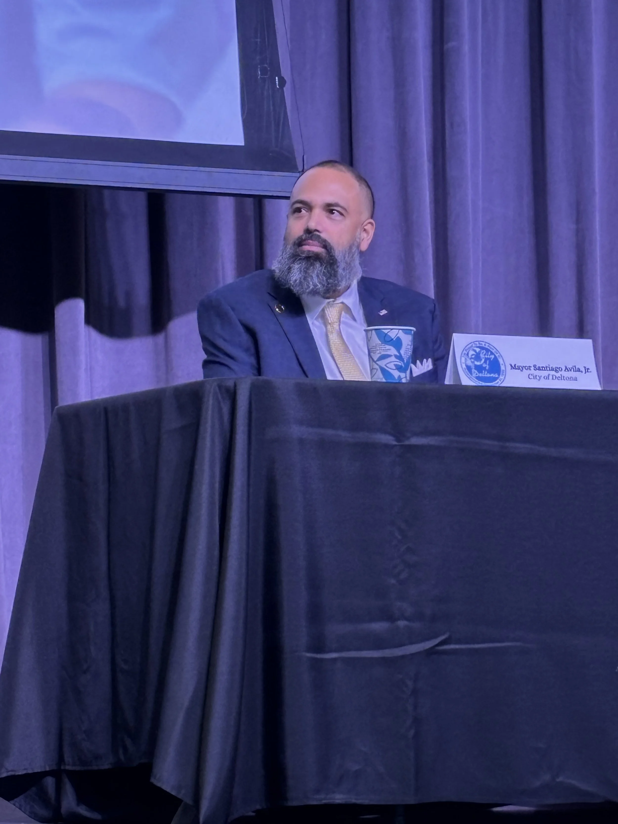 A man with a beard and mustache sitting at a conference table with a nameplate that says "Mayor Santiago Avila, Jr., City of Deltona." He is wearing a dark suit, white shirt, and a yellow tie, and is looking to his right. There is a large screen behind him displaying a presentation, and purple curtains cover the background.