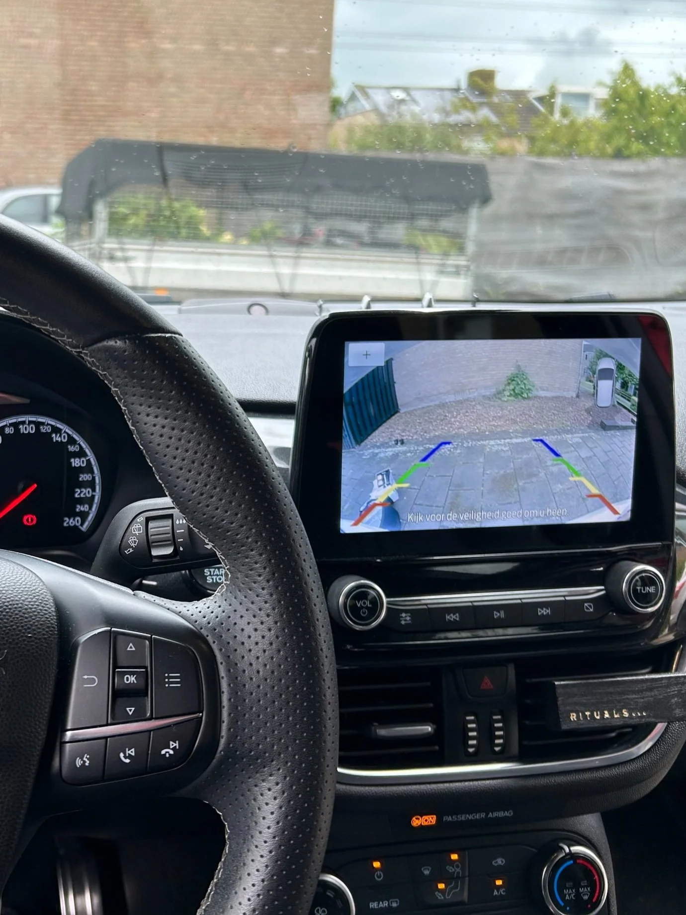 Binnenkant van een auto met een dashboard, stuur en een achteruitkijkcamera display. Het display toont een achteruitrijcamera met gekleurde lijnen voor de parkeerhulp.