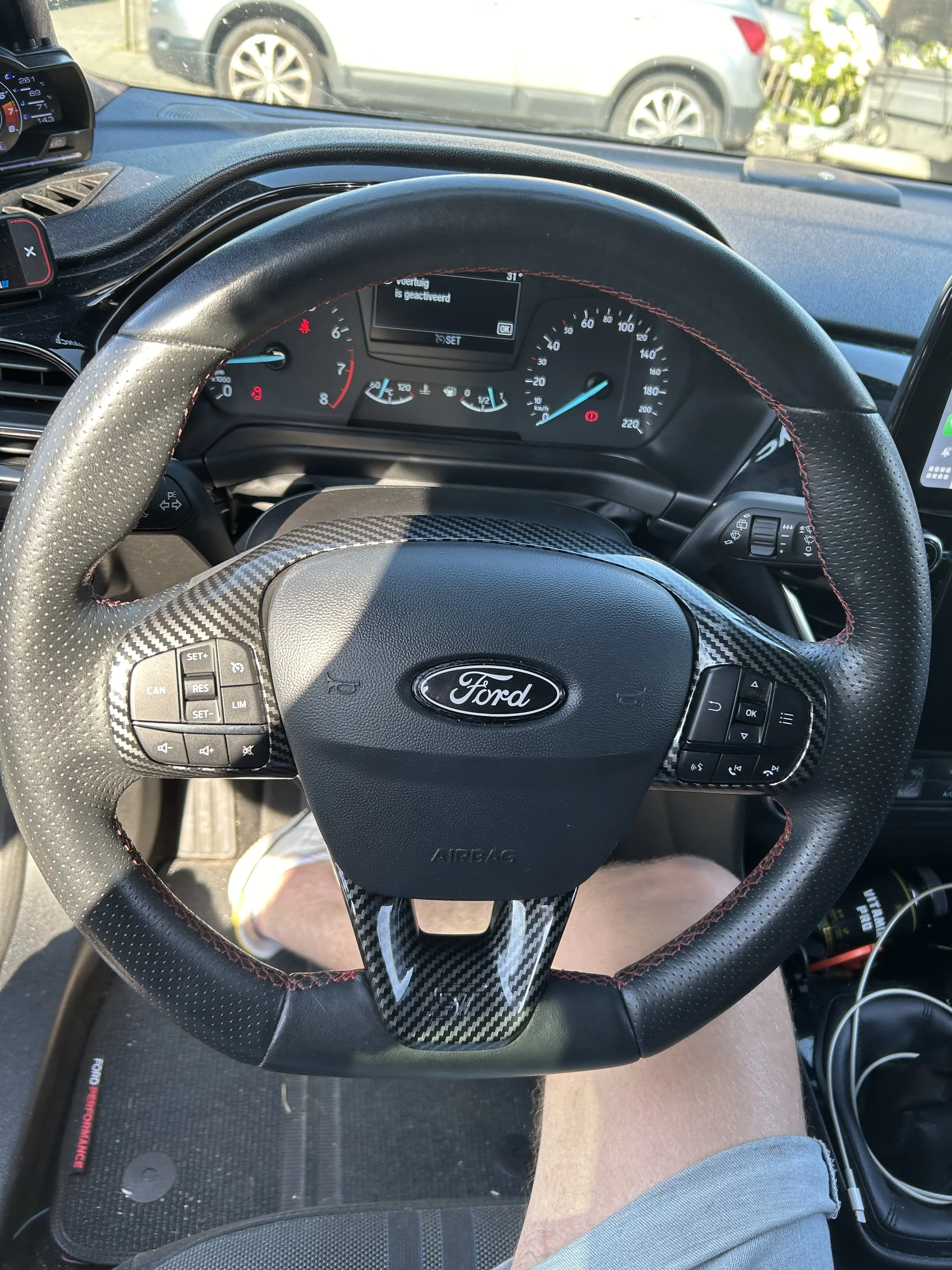 Binnenkant van een auto met een stuurwiel van een Ford, snelheidsmeter, brandstofmeter, en een digitale display op de achtergrond, met een auto en bloemen buiten zichtbaar. Voorzien van cruise control.