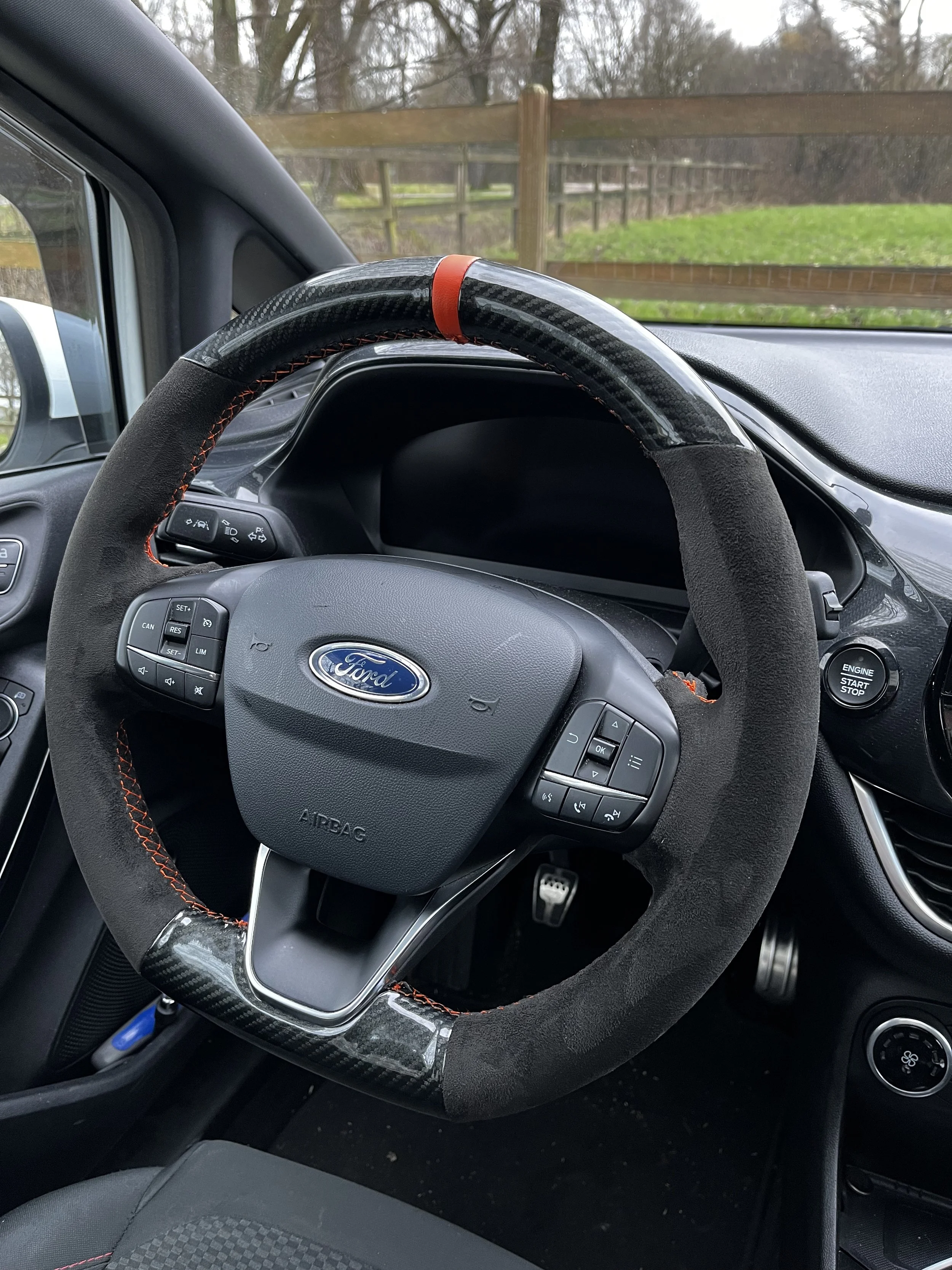 Interieur van een auto met een stuurwiel dat is bedekt met een sportief stuurwielhoesje, een Ford logo op het stuur en een startknop aan de rechterkant. Voorzien van cruise control.